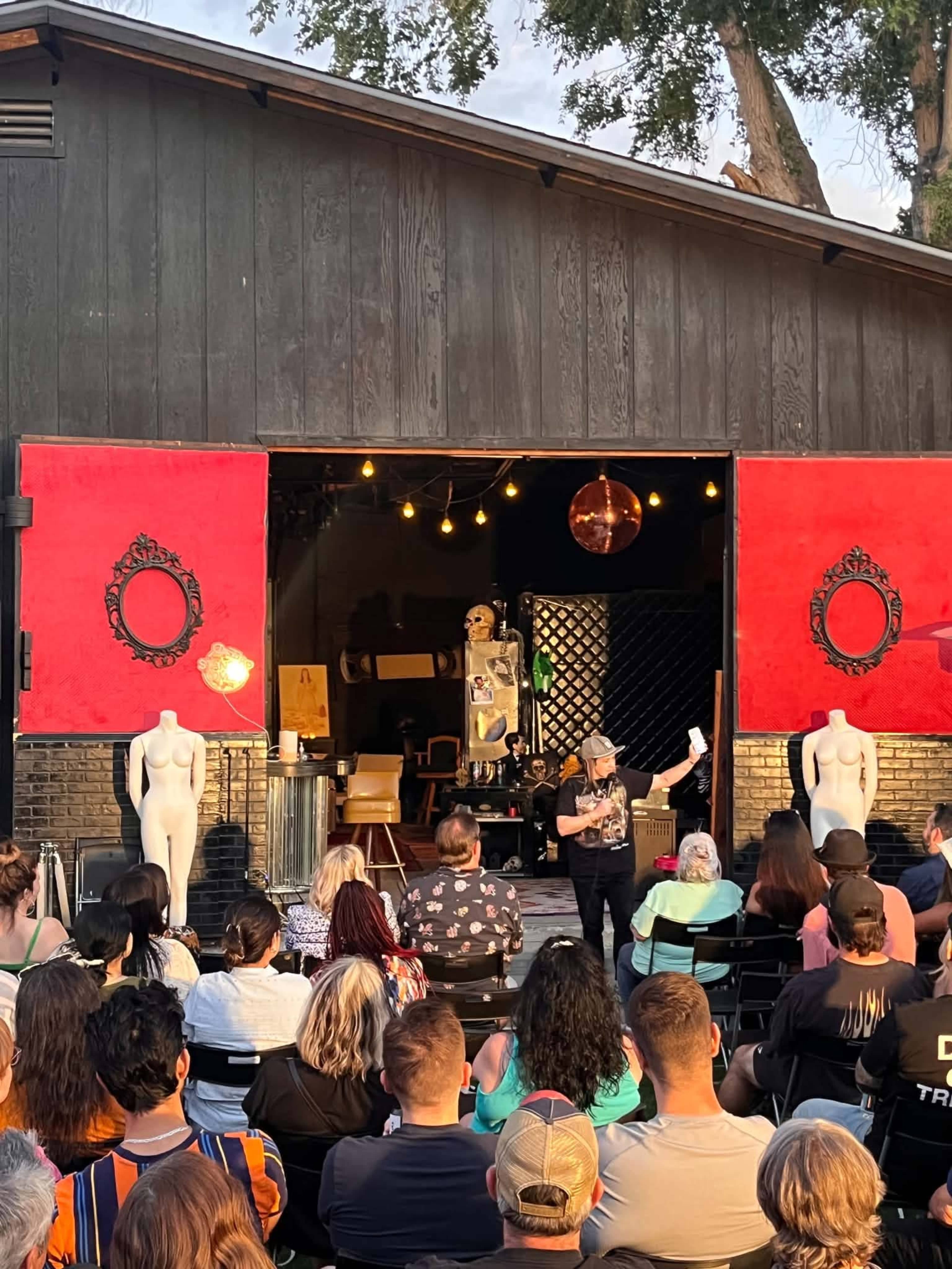 A speaker stands in front of an audience inside a venue with bright red doors, flanked by mannequin figures, while decorative lights illuminate the space.