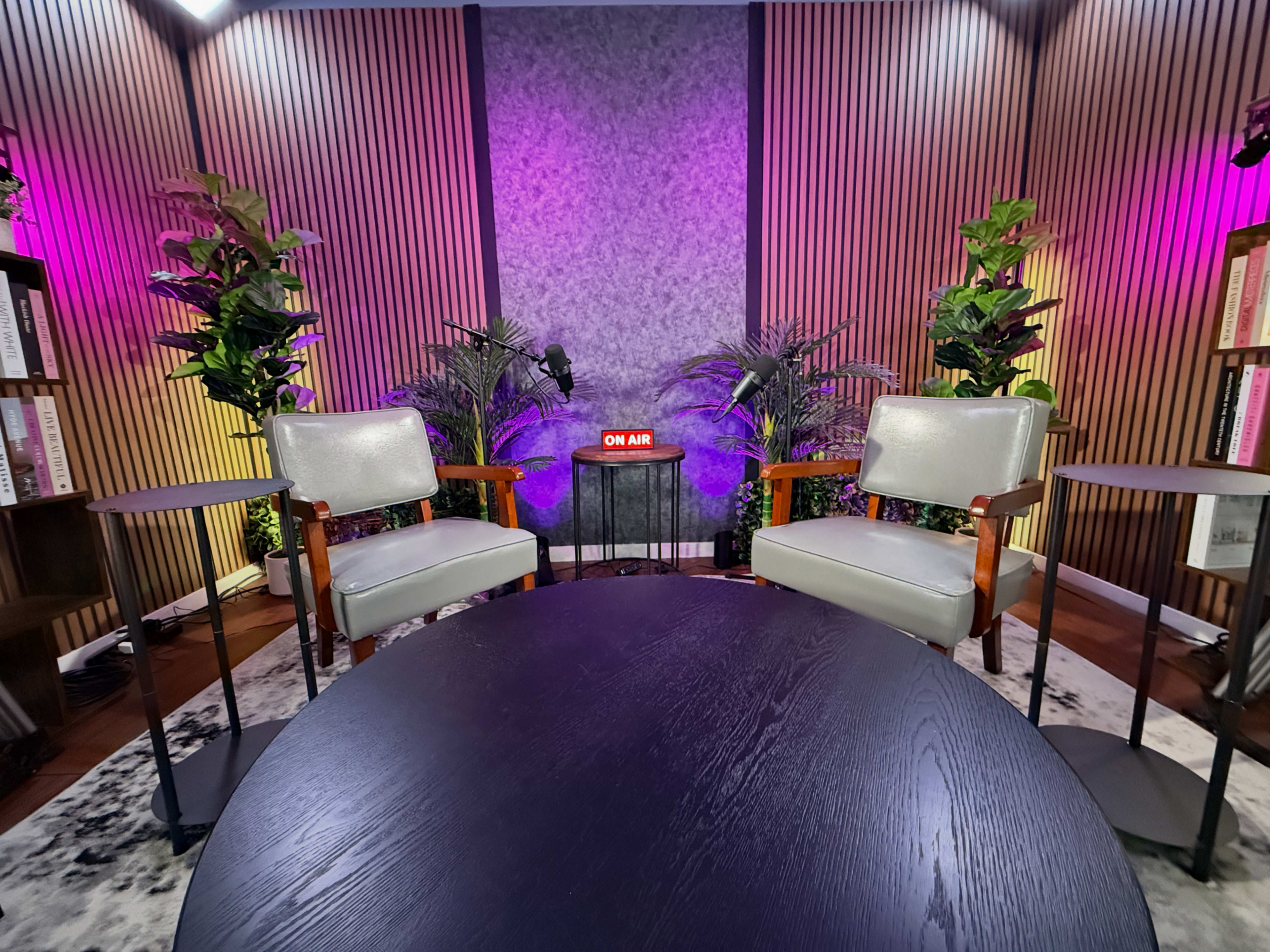 A modern studio setup features two gray chairs facing a black round table, surrounded by plants and illuminated with purple lighting, with an "ON AIR" sign prominently displayed.