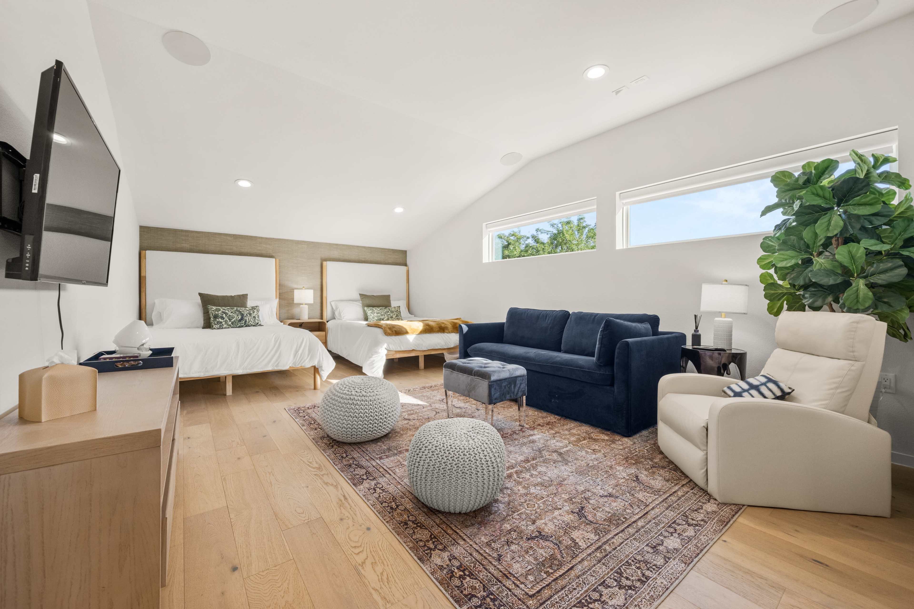 The image shows a modern living space with two beds, a blue couch, a wooden TV unit, and a decorative rug, all featuring large windows that allow natural light.