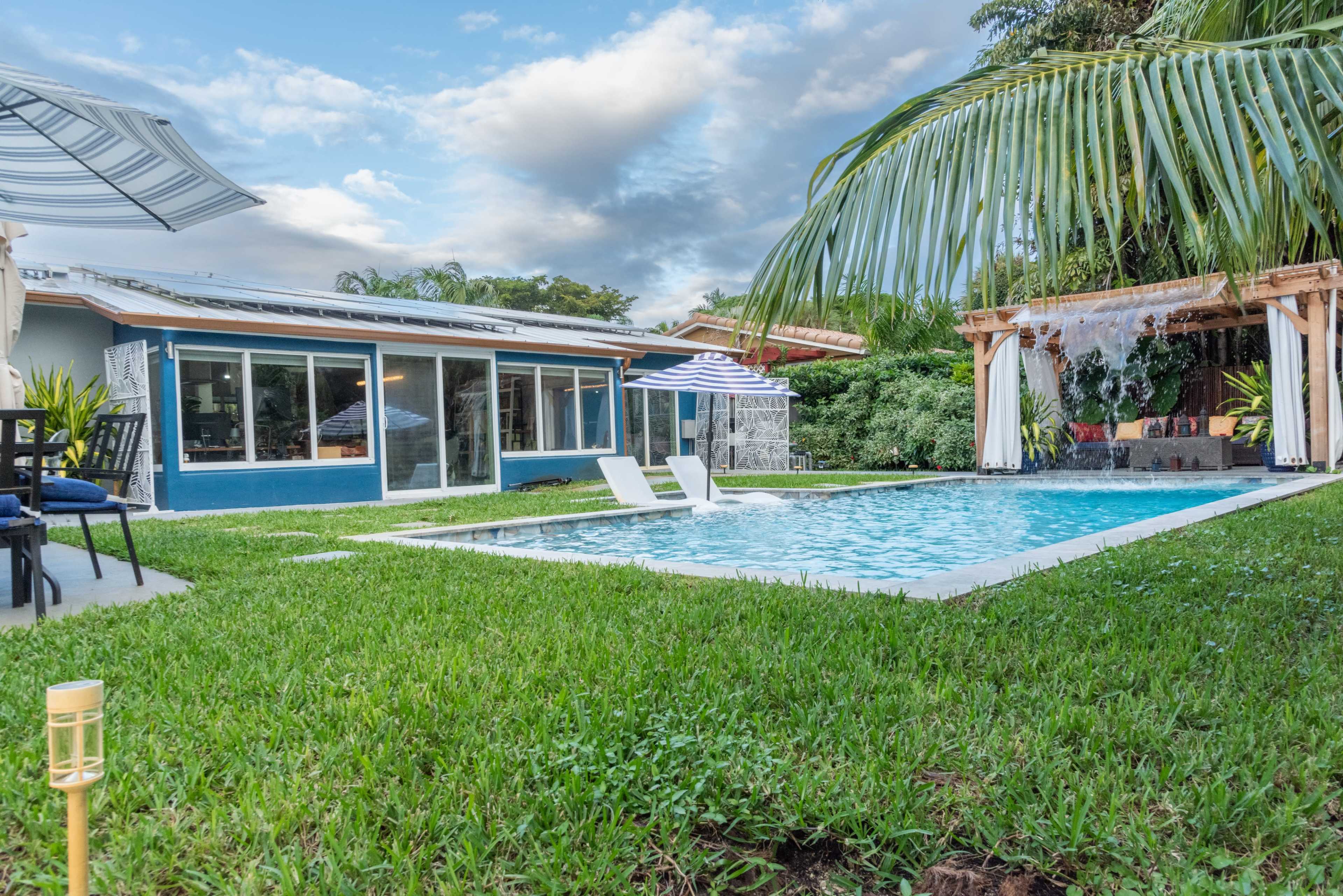 The image shows a modern house with large windows beside a swimming pool, surrounded by a well-maintained lawn and tropical plants.