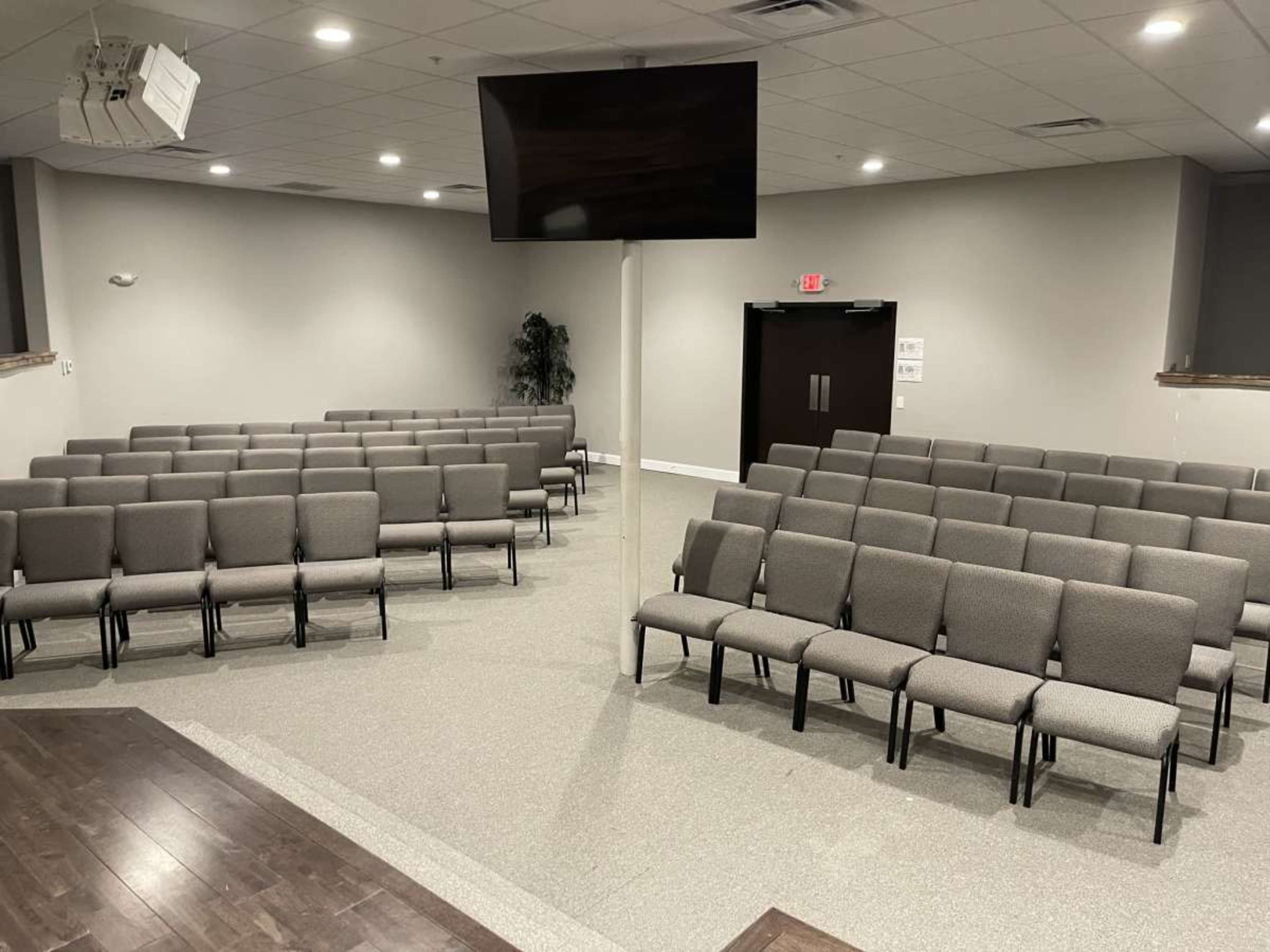 The image shows a spacious, empty meeting room with several rows of gray chairs arranged in a semi-circle facing a central podium and a large screen above.
