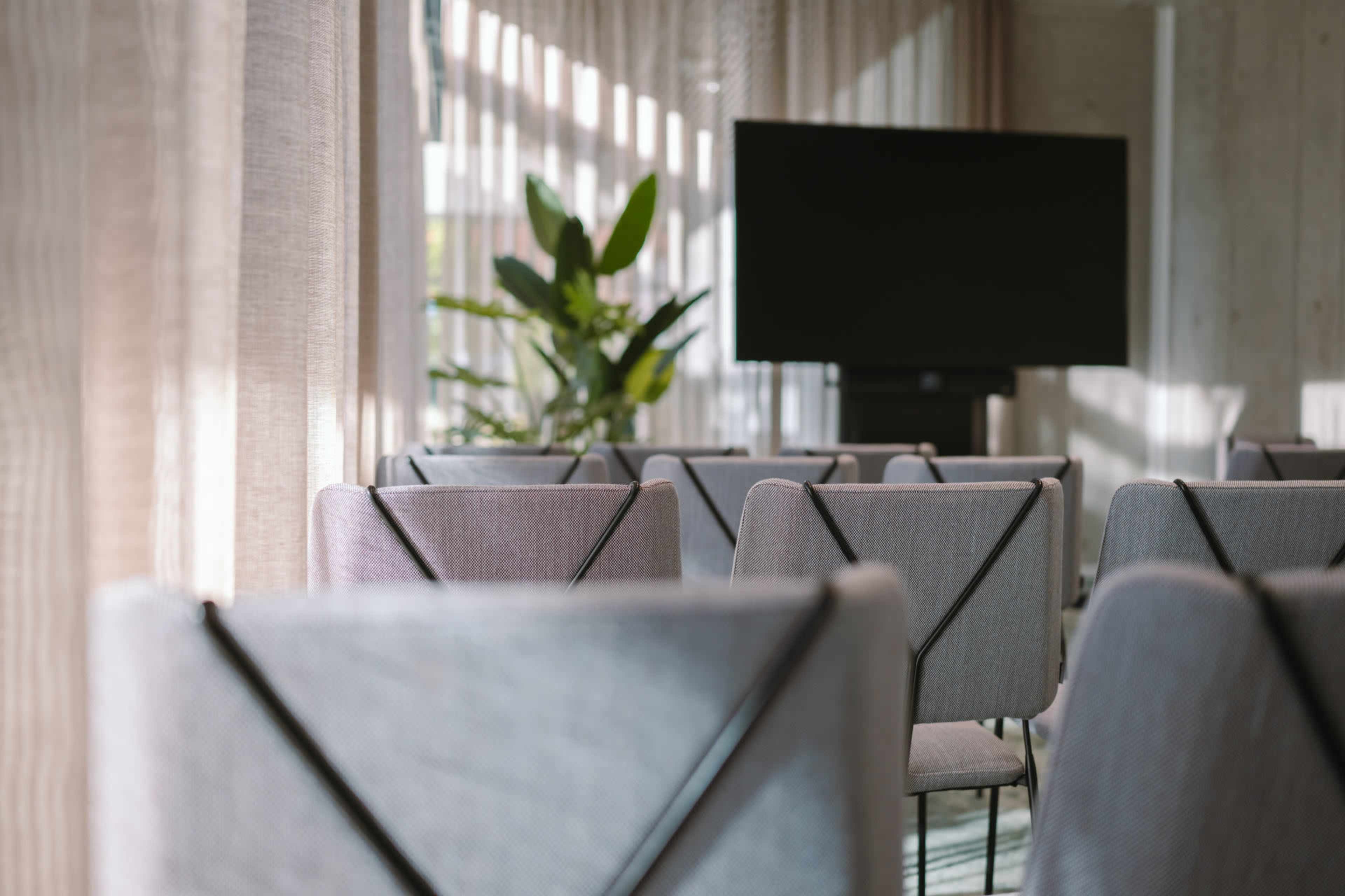 A row of gray chairs is positioned in a well-lit room with sheer curtains and a large screen at the front.