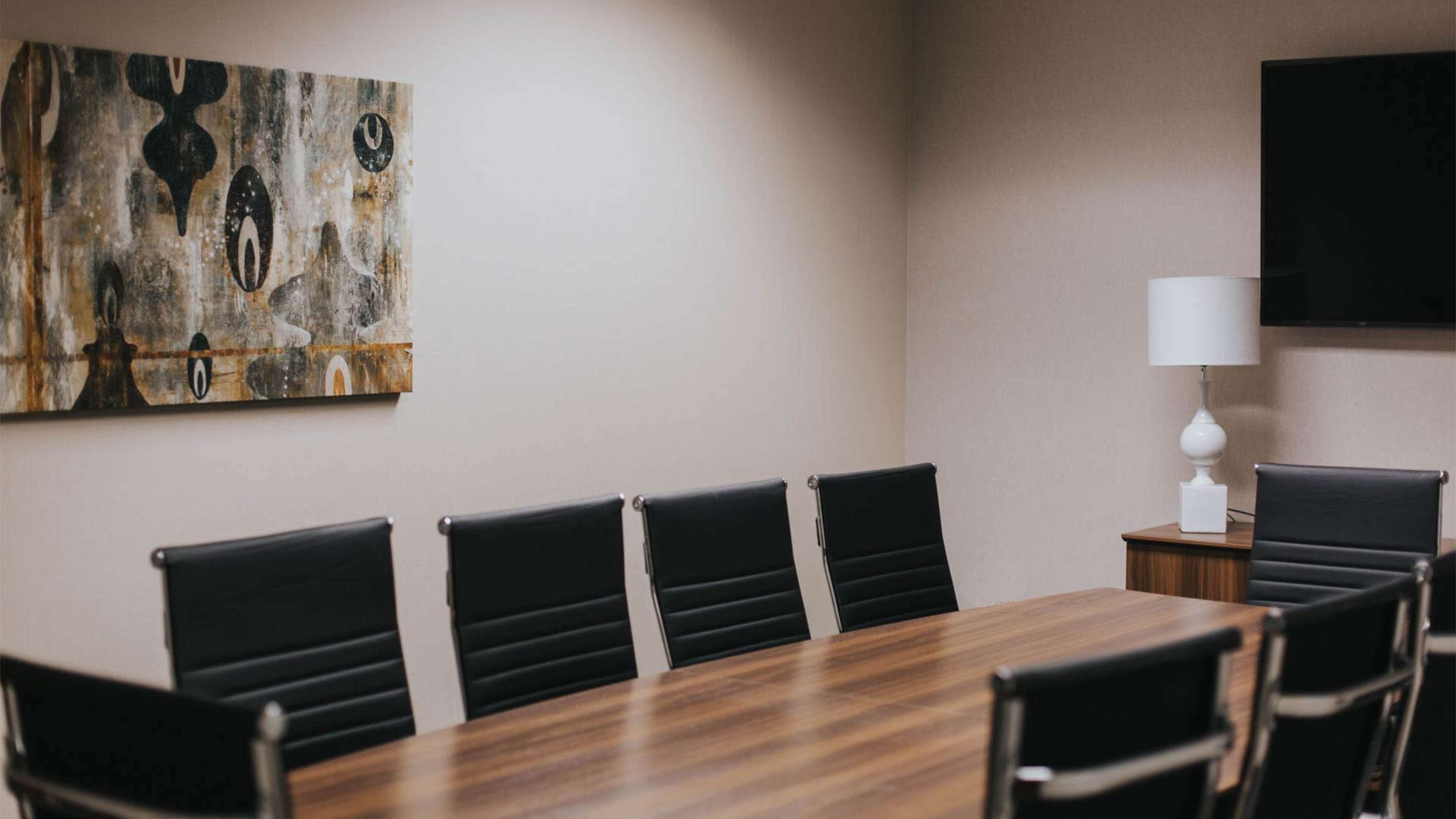 A modern conference room features a large wooden table surrounded by black leather chairs, with a wall-mounted TV and abstract artwork decorating the space.