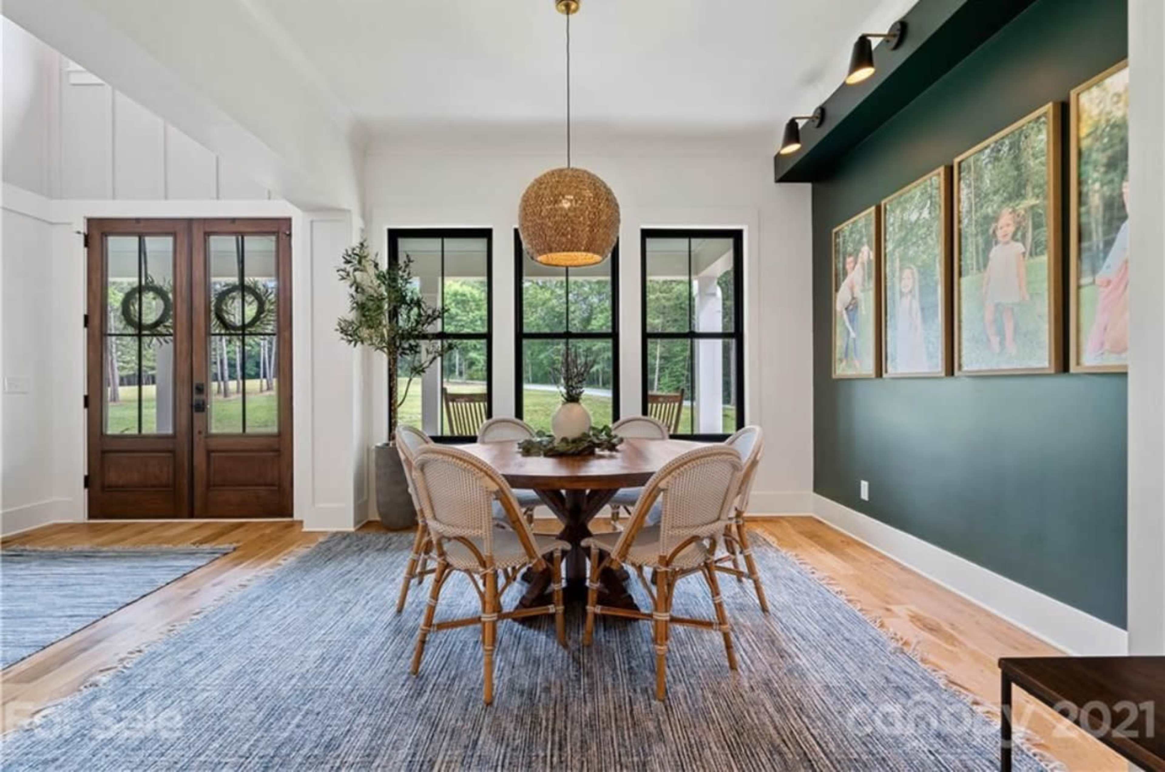 A dining area features a round table surrounded by six chairs, with large windows and framed photos on the wall.