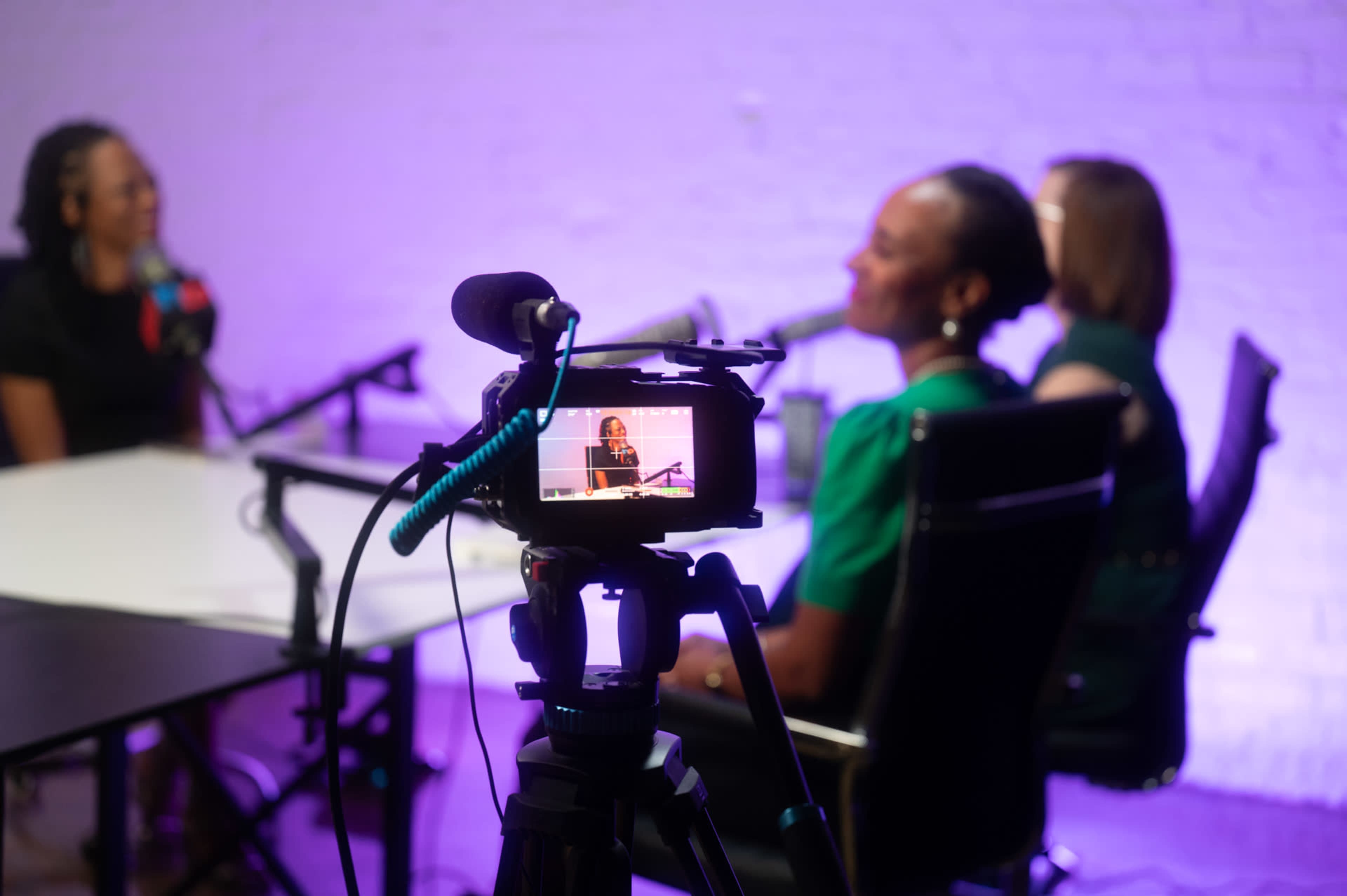 The image shows a camera focused on two women engaged in a discussion while seated at a table, with microphones and soft purple lighting in the background.