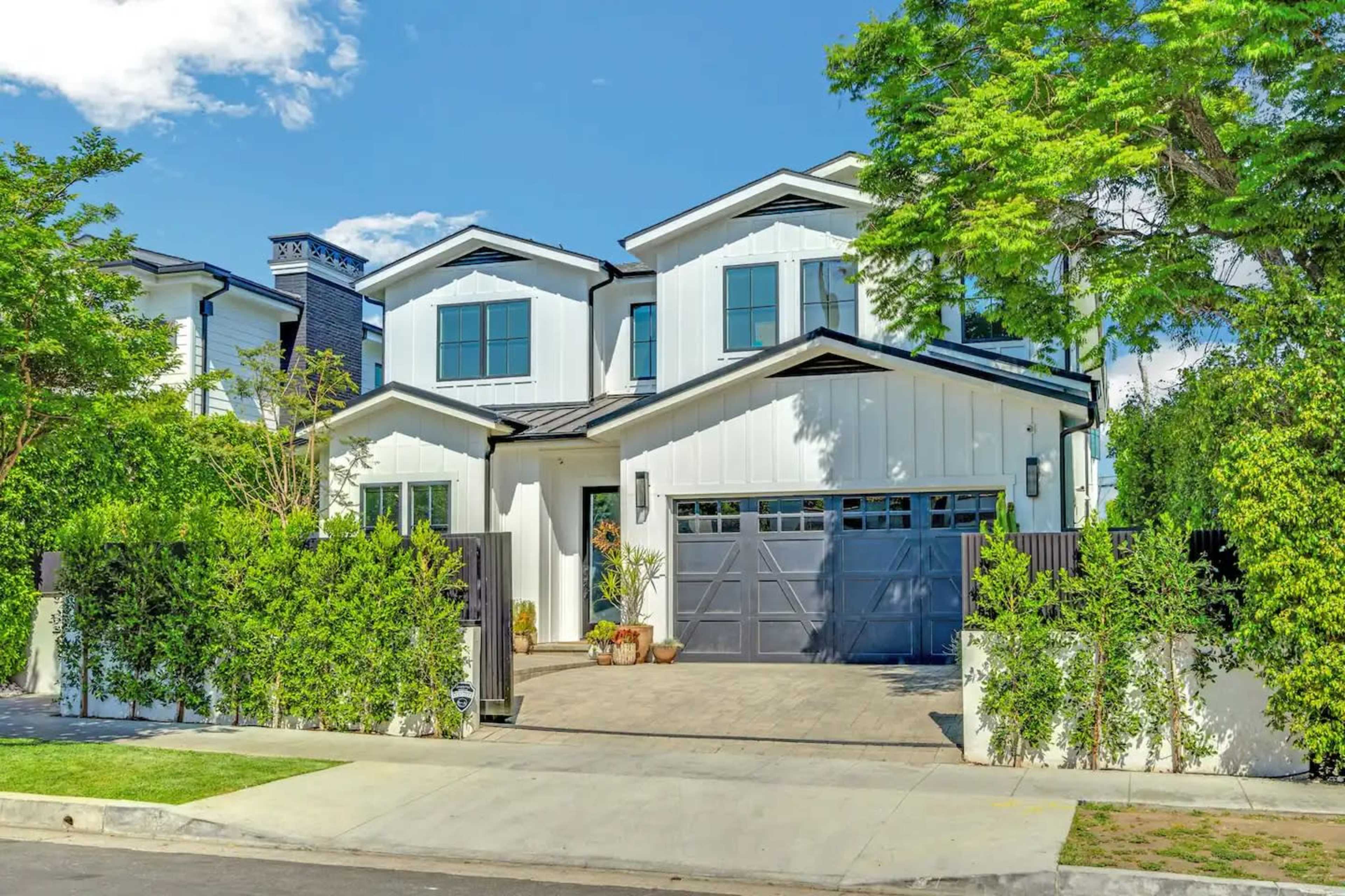 A white two-story house with a black roof and a paved driveway is surrounded by greenery and features a two-car garage.