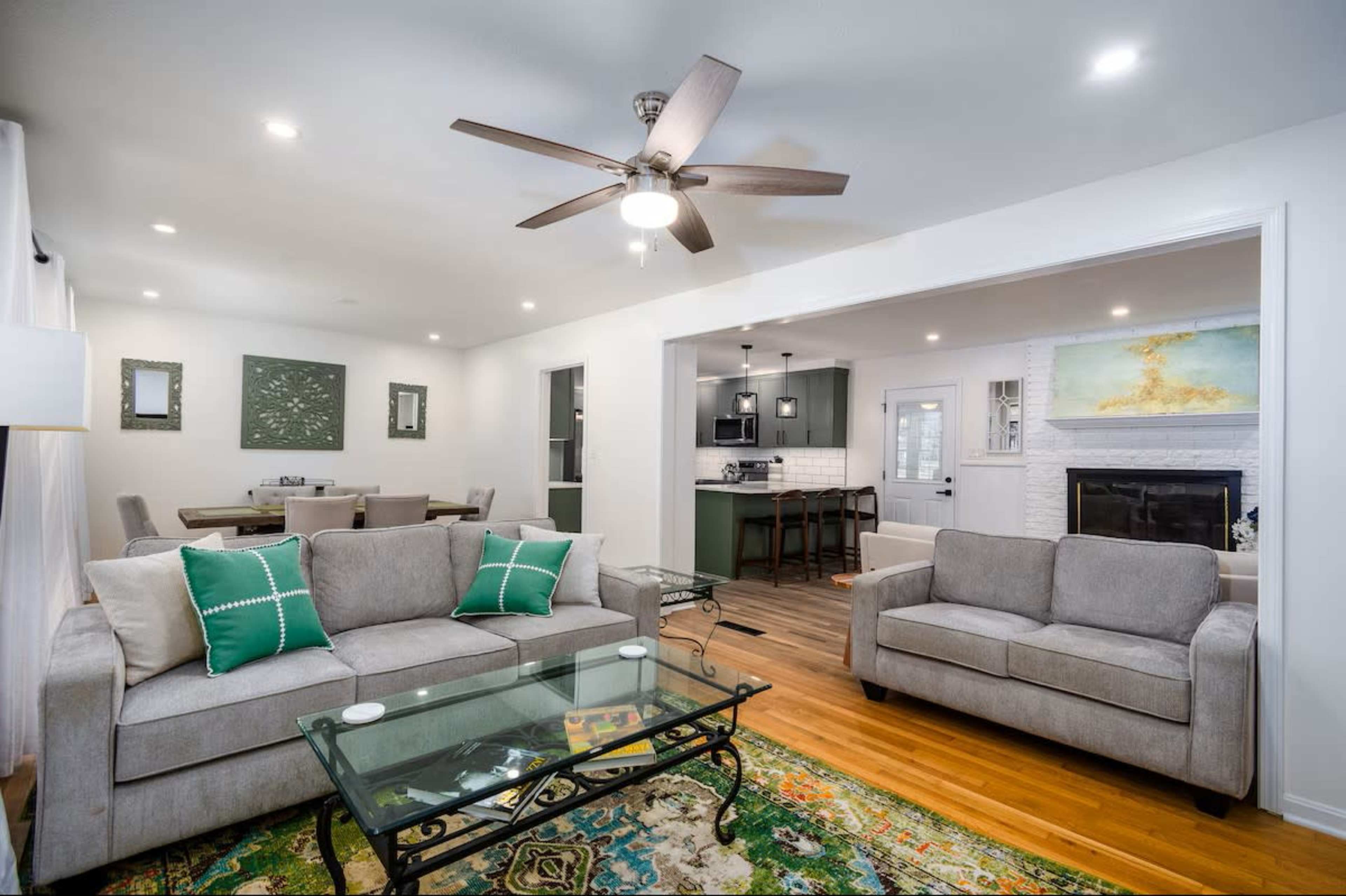The image shows a living room with a gray sofa and loveseat facing a glass coffee table, a ceiling fan overhead, and an open layout leading to a dining area and kitchen.