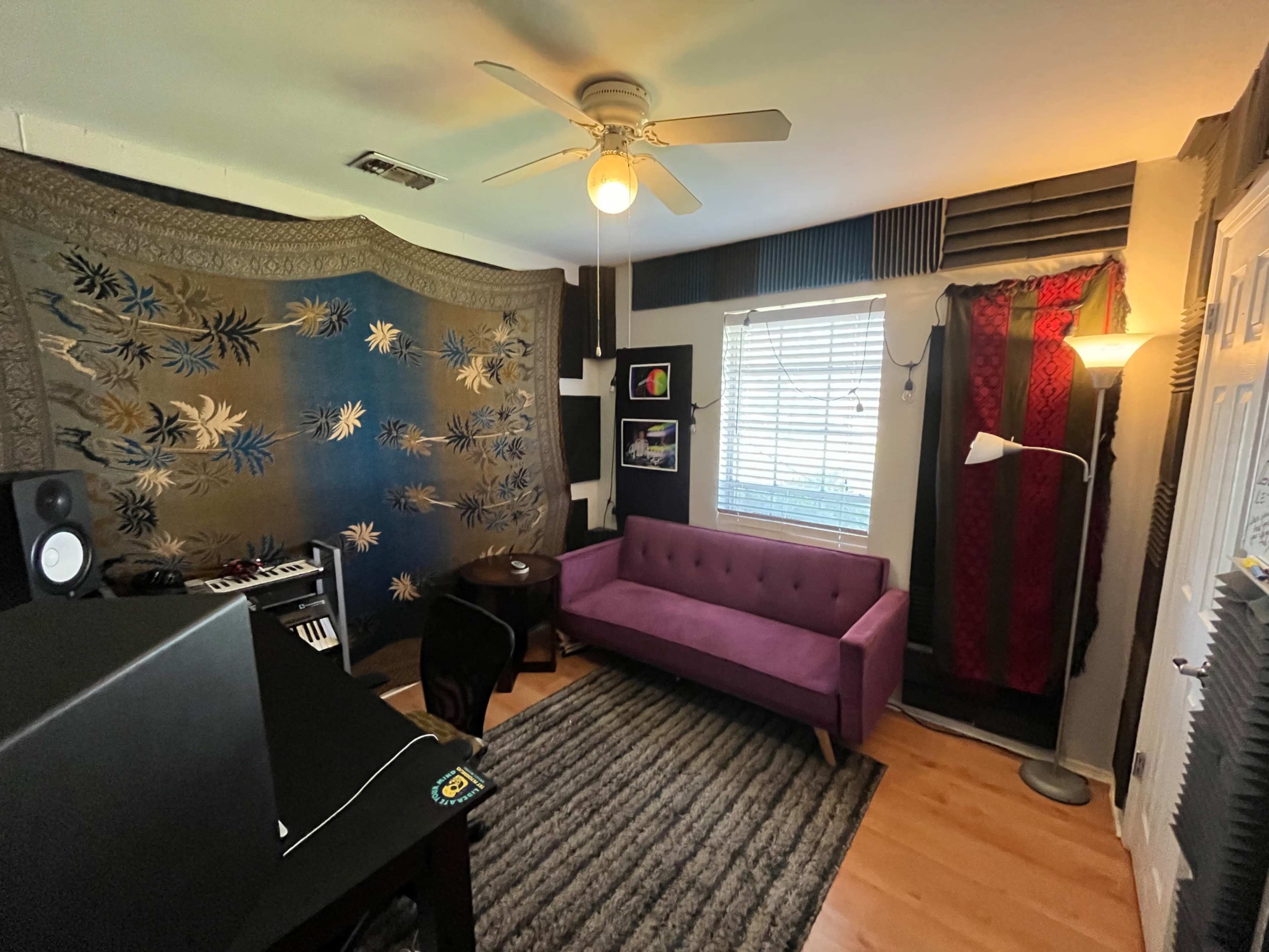 The image shows a small room featuring a purple sofa, a black desk with speakers, and decorative wall hangings.