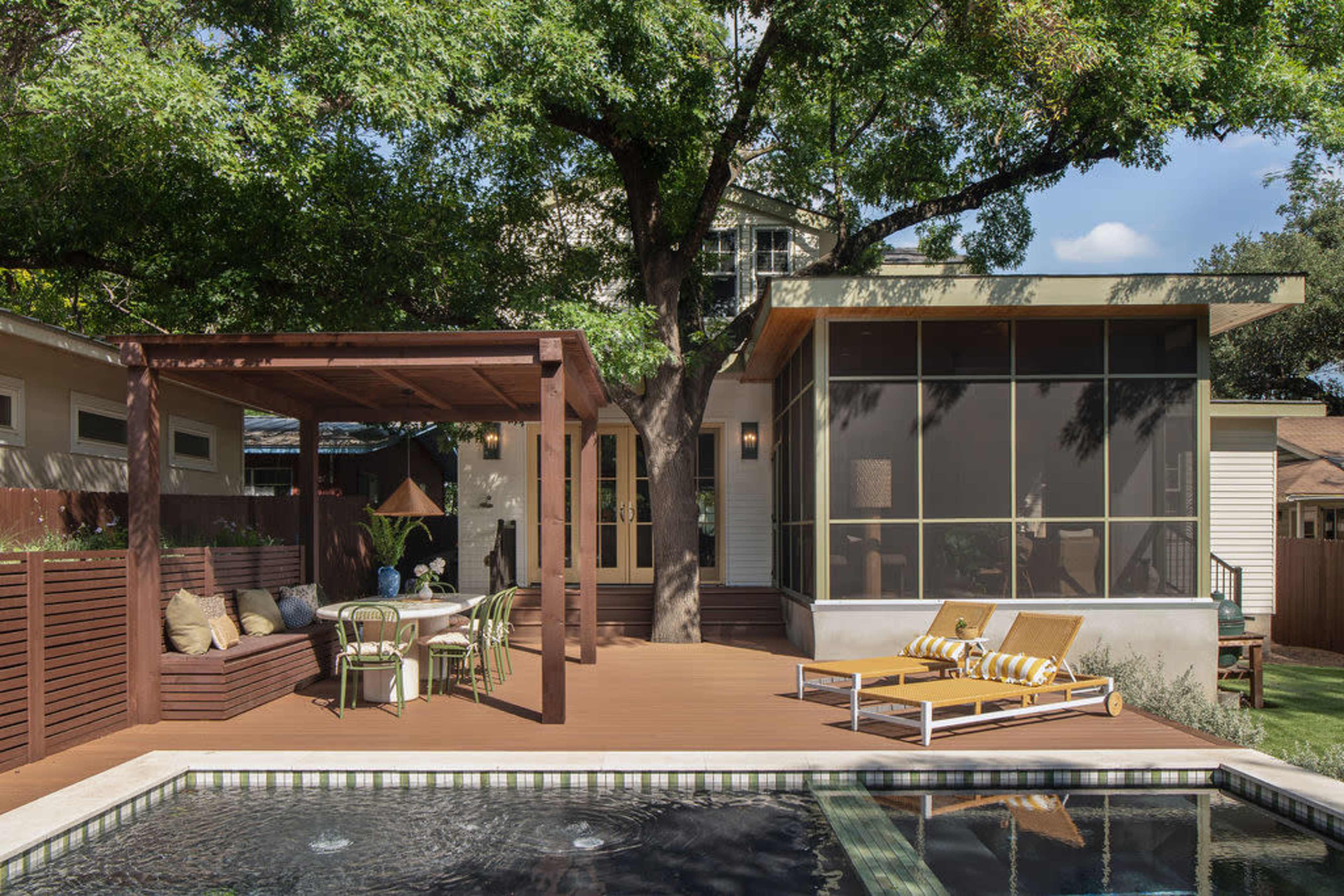 The image shows a backyard patio featuring a wooden deck with a pool, lounging chairs, a shaded seating area, and a house with large windows.