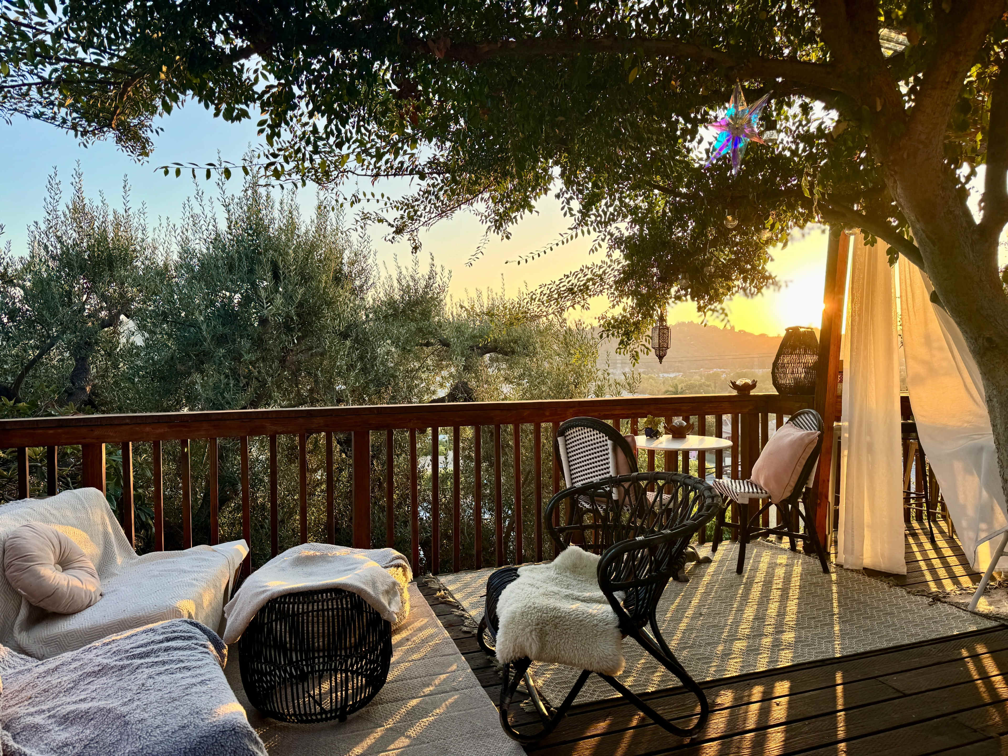 A deck overlooking a landscape is adorned with chairs, cushions, and plants as the sun sets in the background.