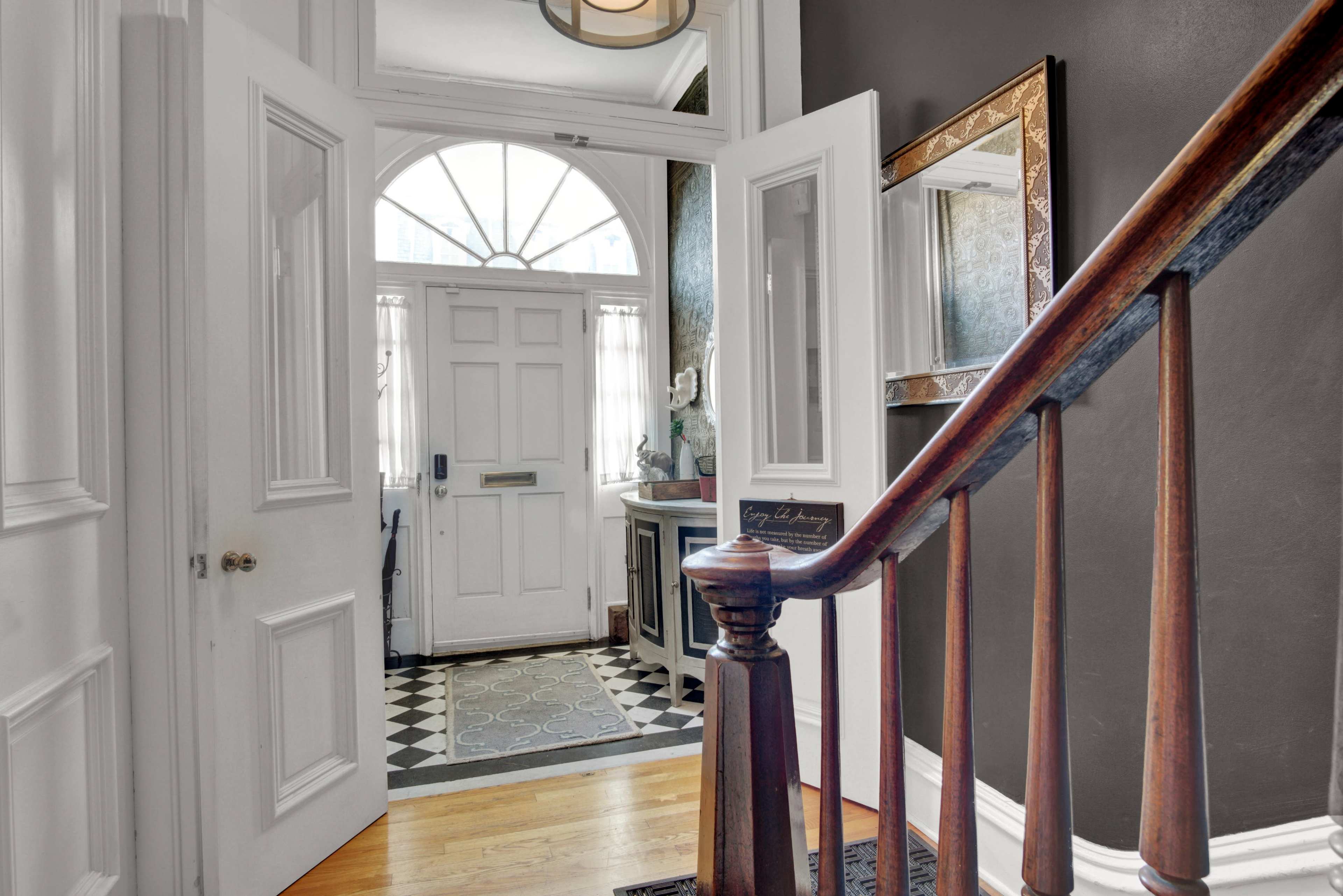 The image shows a hallway entrance with a staircase leading to an open front door, revealing a patterned floor and a glimpse of light in the entryway.