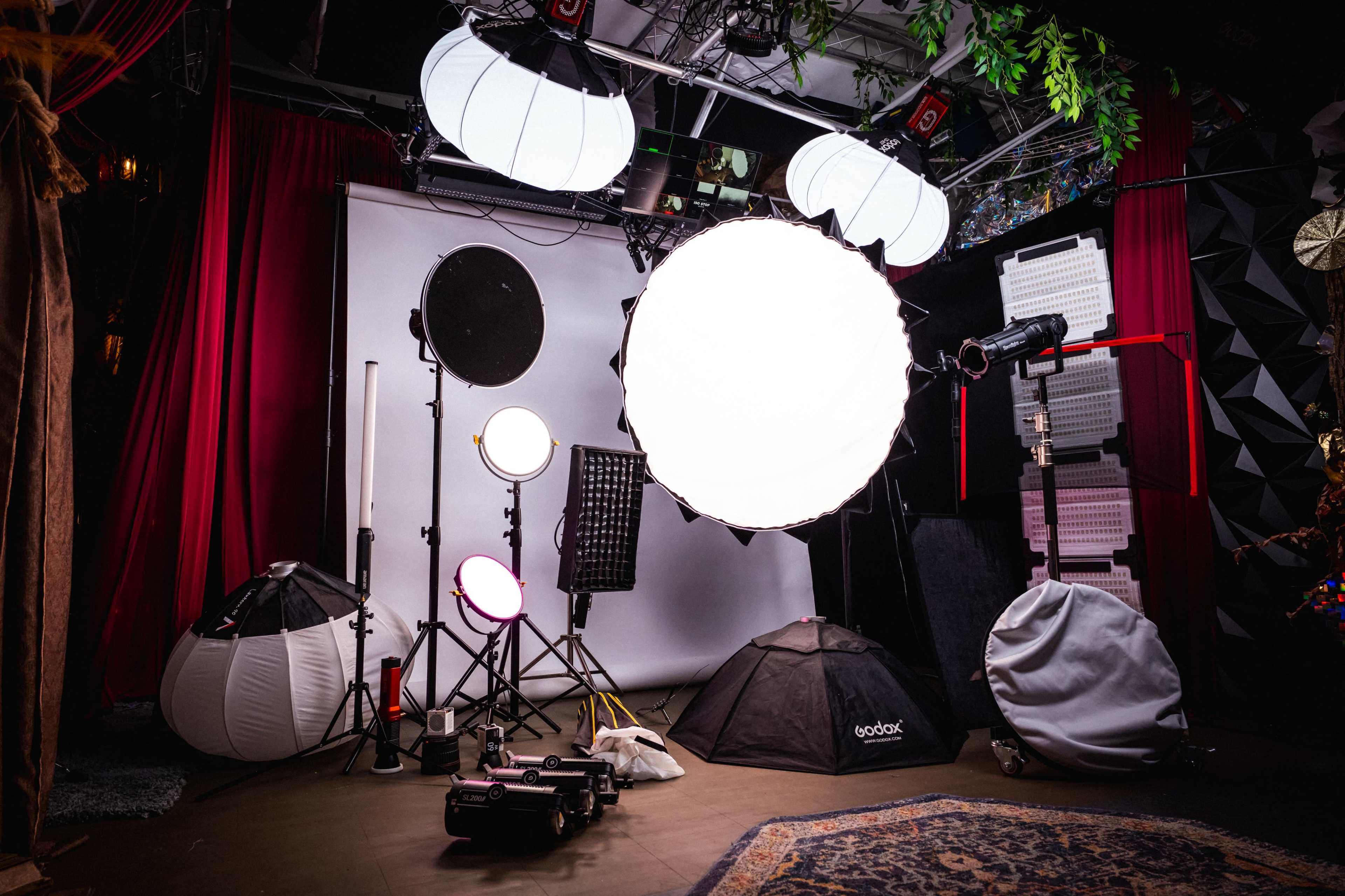 The image shows a photography studio equipped with various lighting setups, including softboxes and a boom arm, surrounded by portable light stands and backdrops.