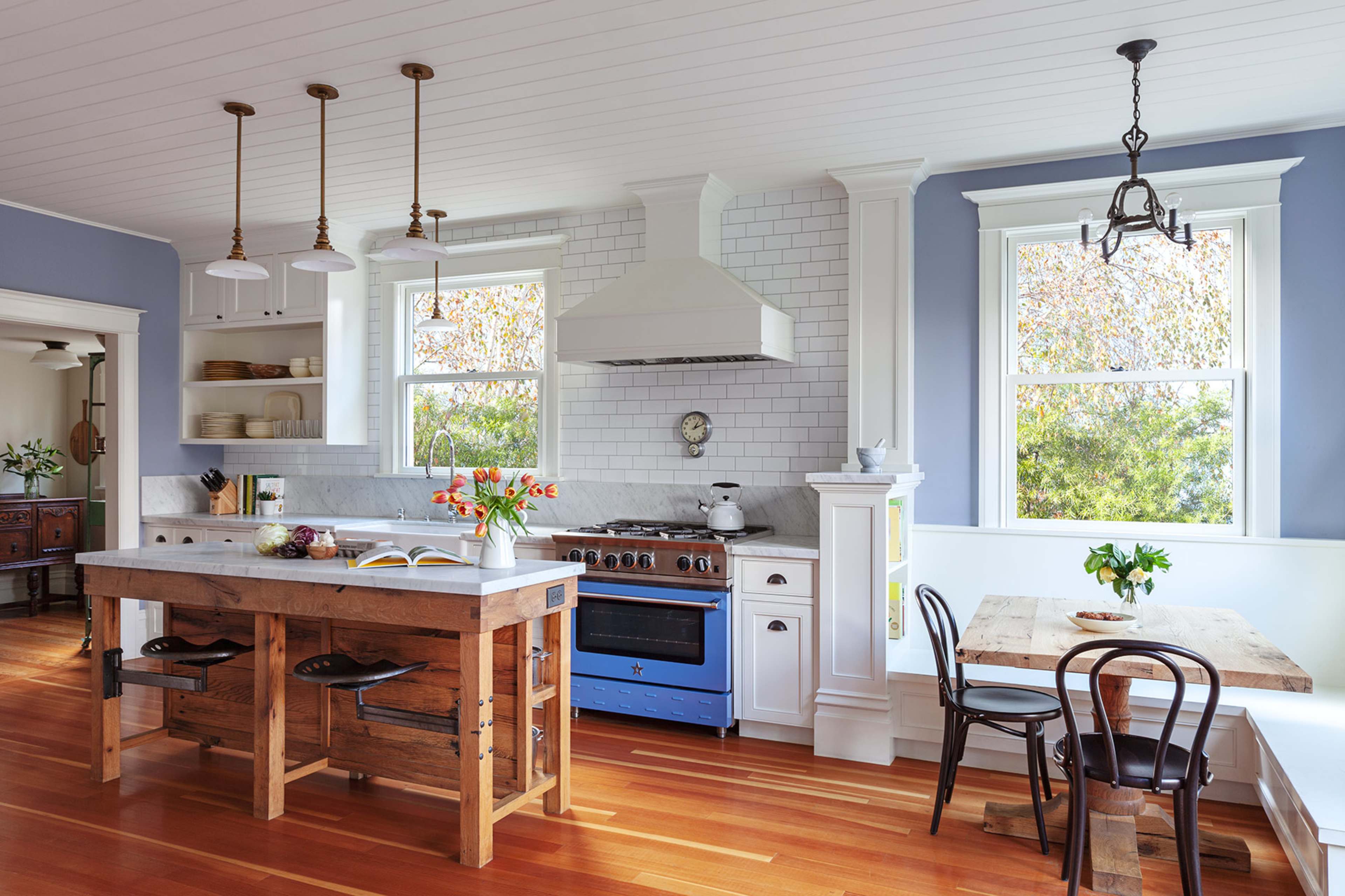 The image shows a bright kitchen featuring a wooden island, blue stove, white cabinetry, and large windows with views of greenery.