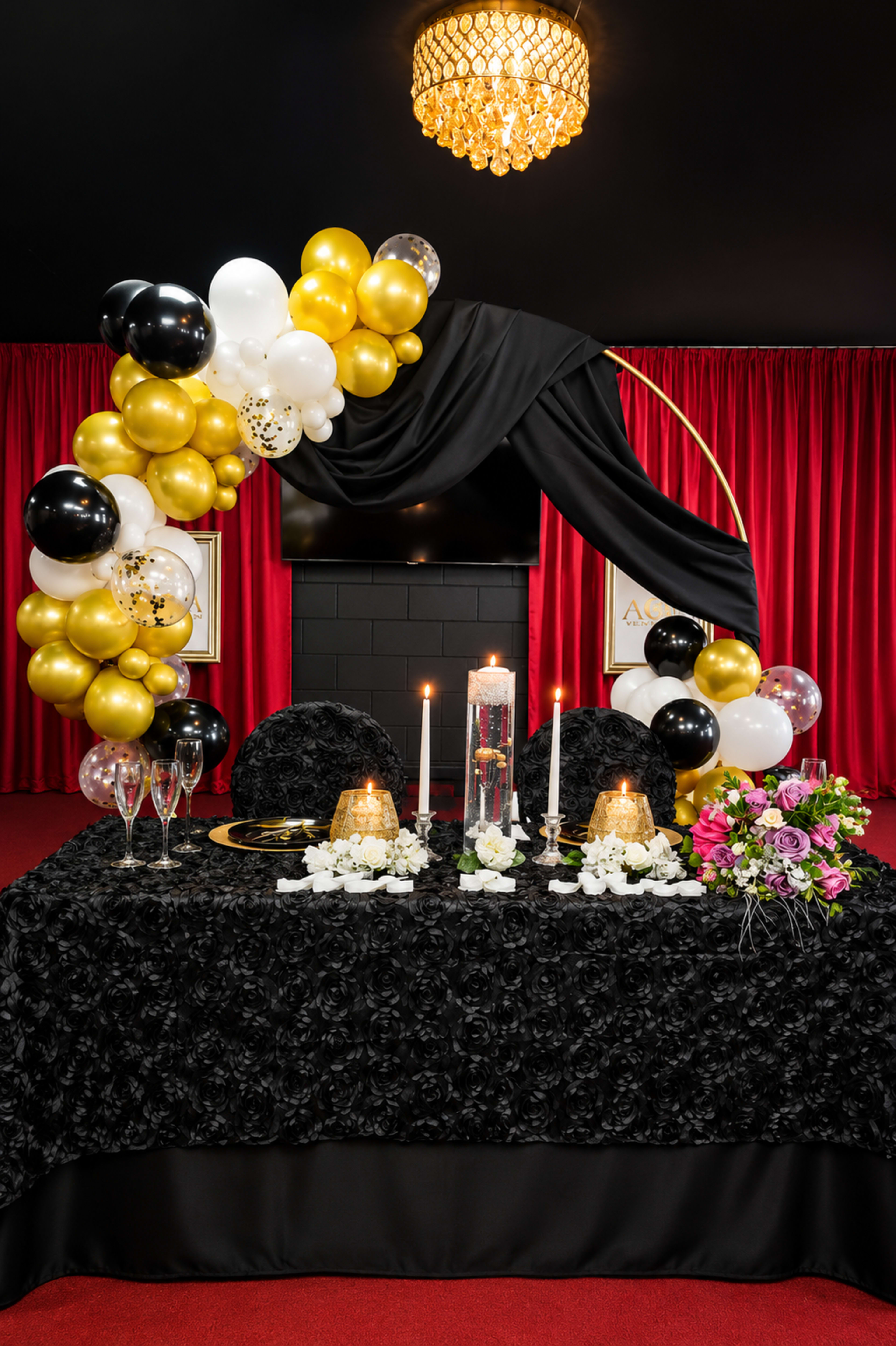 The image features a luxurious party setup with a black table adorned with floral arrangements, candles, gold and black balloons, and a backdrop of red curtains.