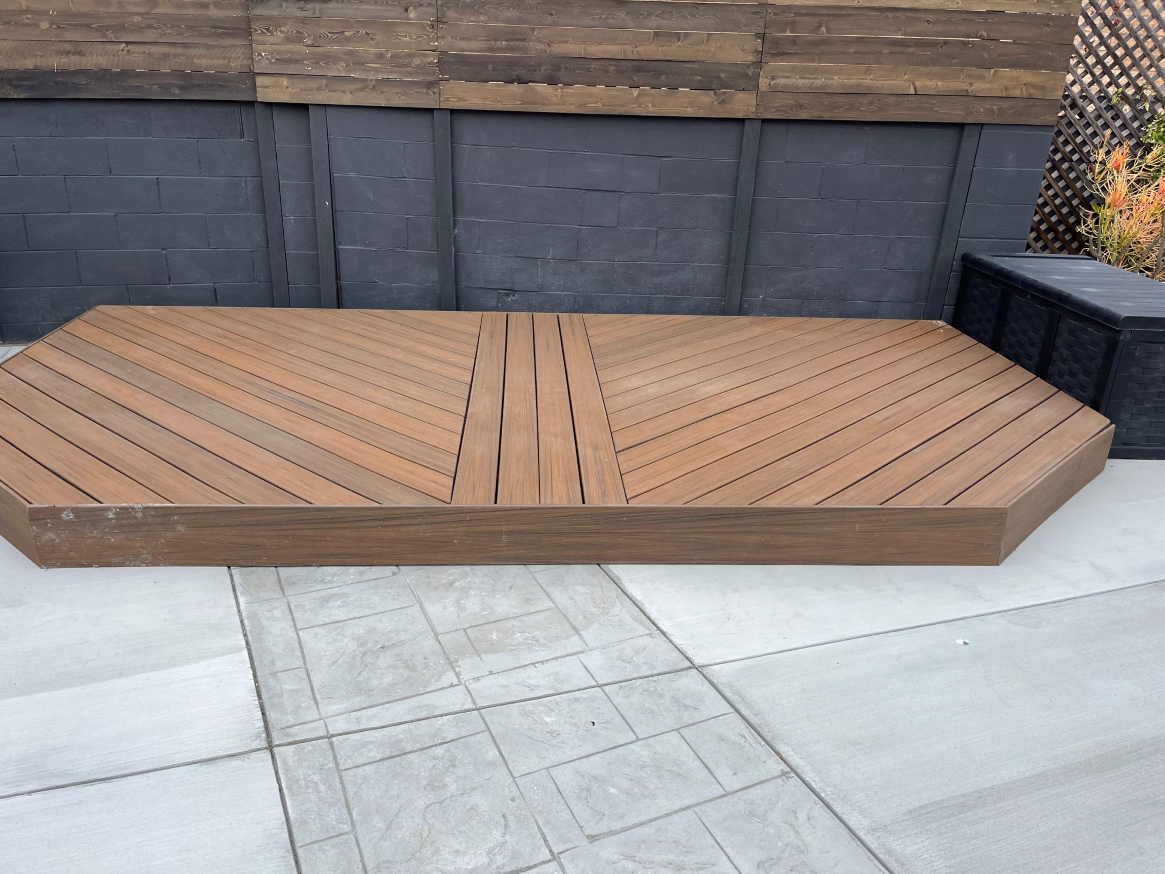 A wooden deck with a hexagonal shape, featuring diagonal planks, positioned next to a paved concrete area.