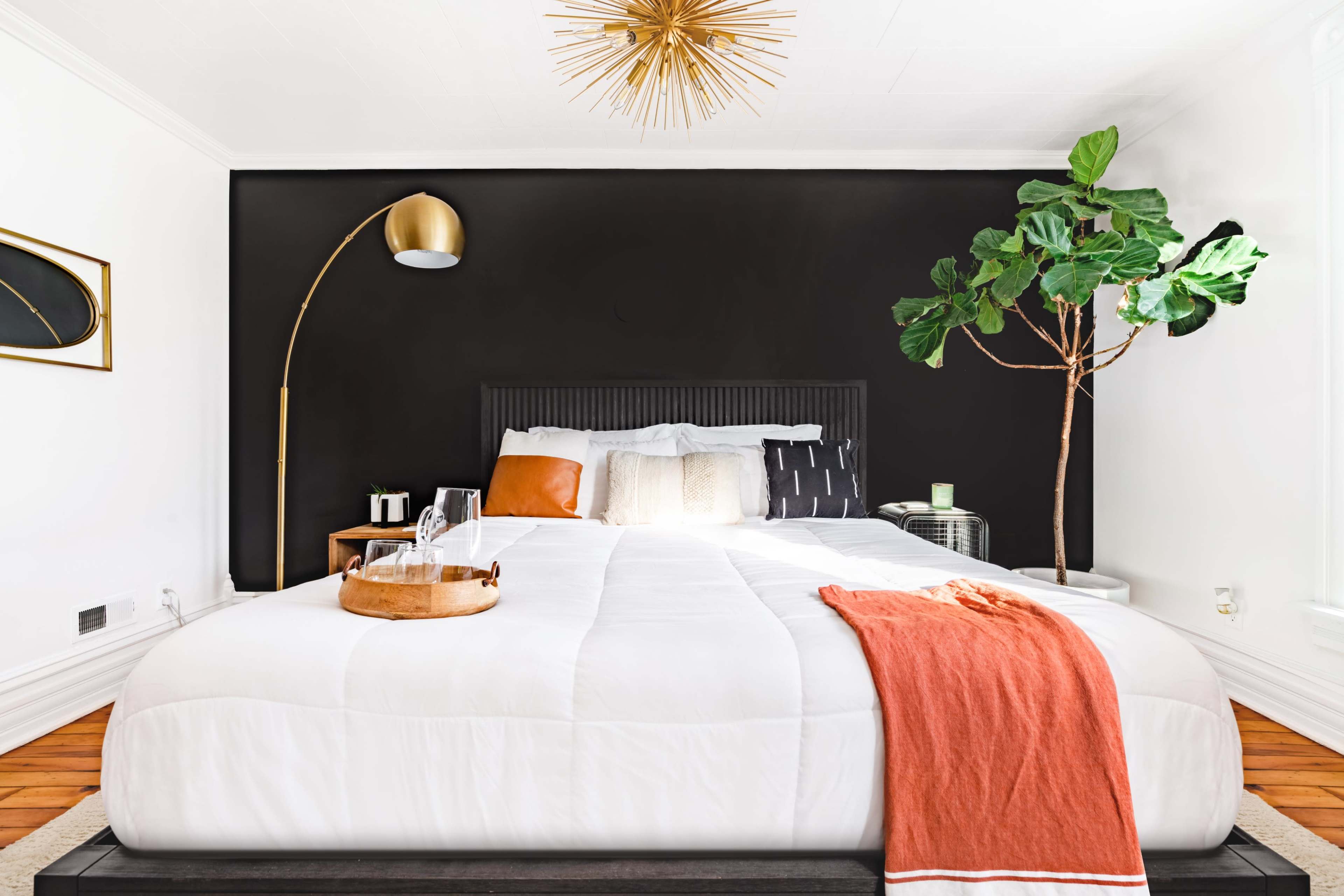 A modern bedroom features a large bed with a white comforter, a gold floor lamp, a decorative tree, and a black accent wall.
