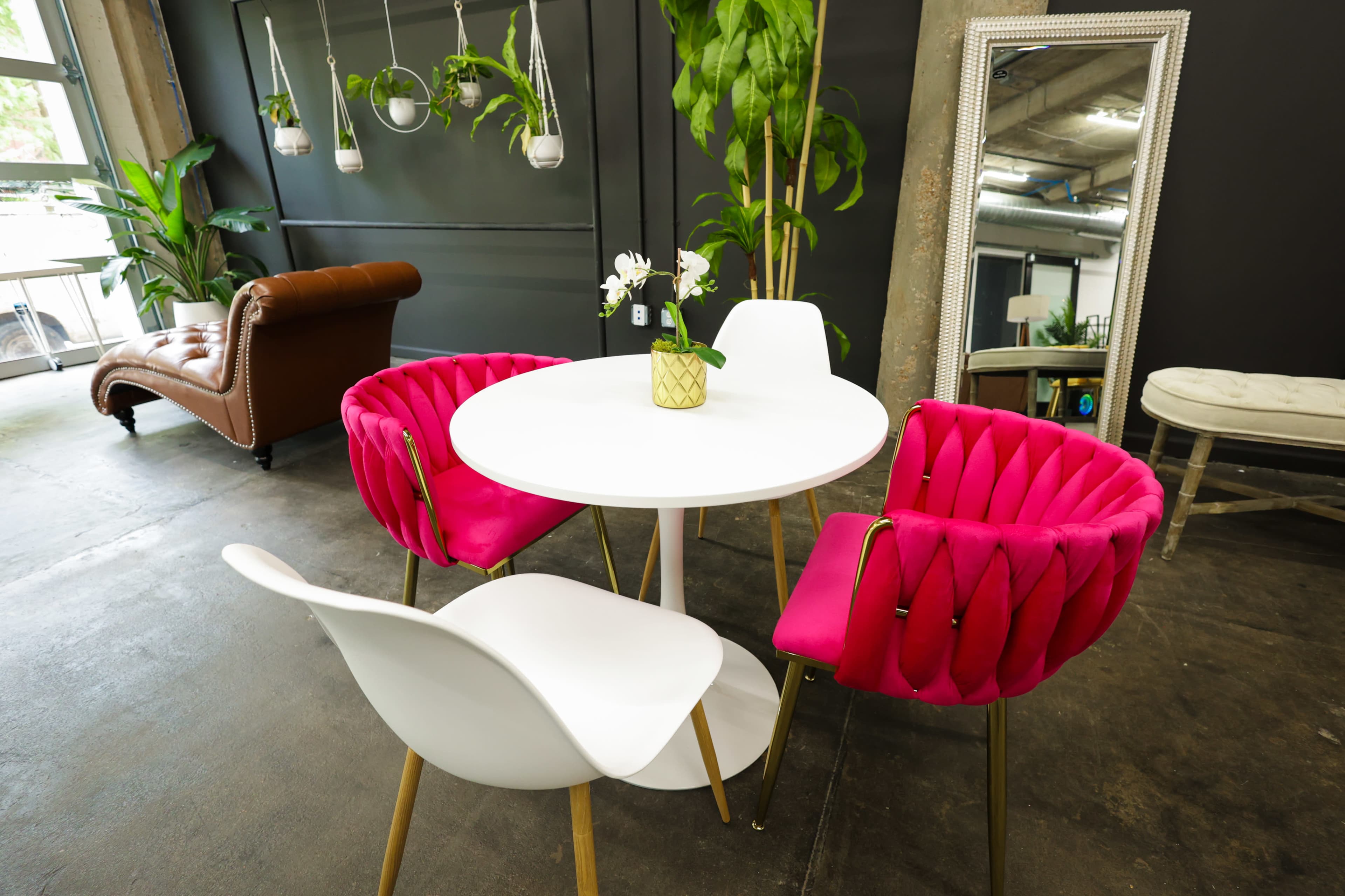 A modern dining area with a round white table surrounded by pink upholstered chairs and a white chair, complemented by plants and a mirror in the background.