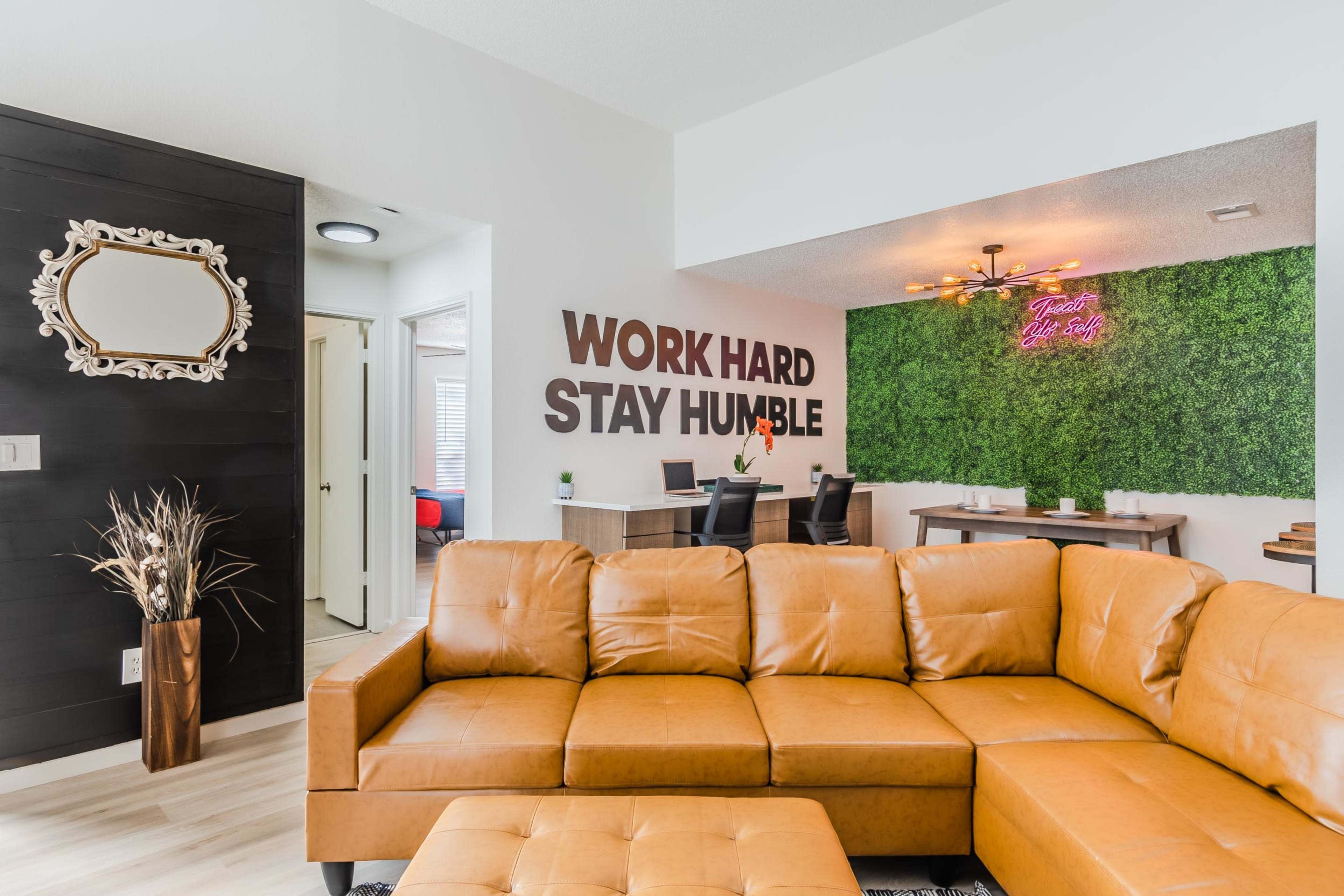A modern living space features a brown leather sectional sofa, with motivational wall art and a green accent wall in the dining area.