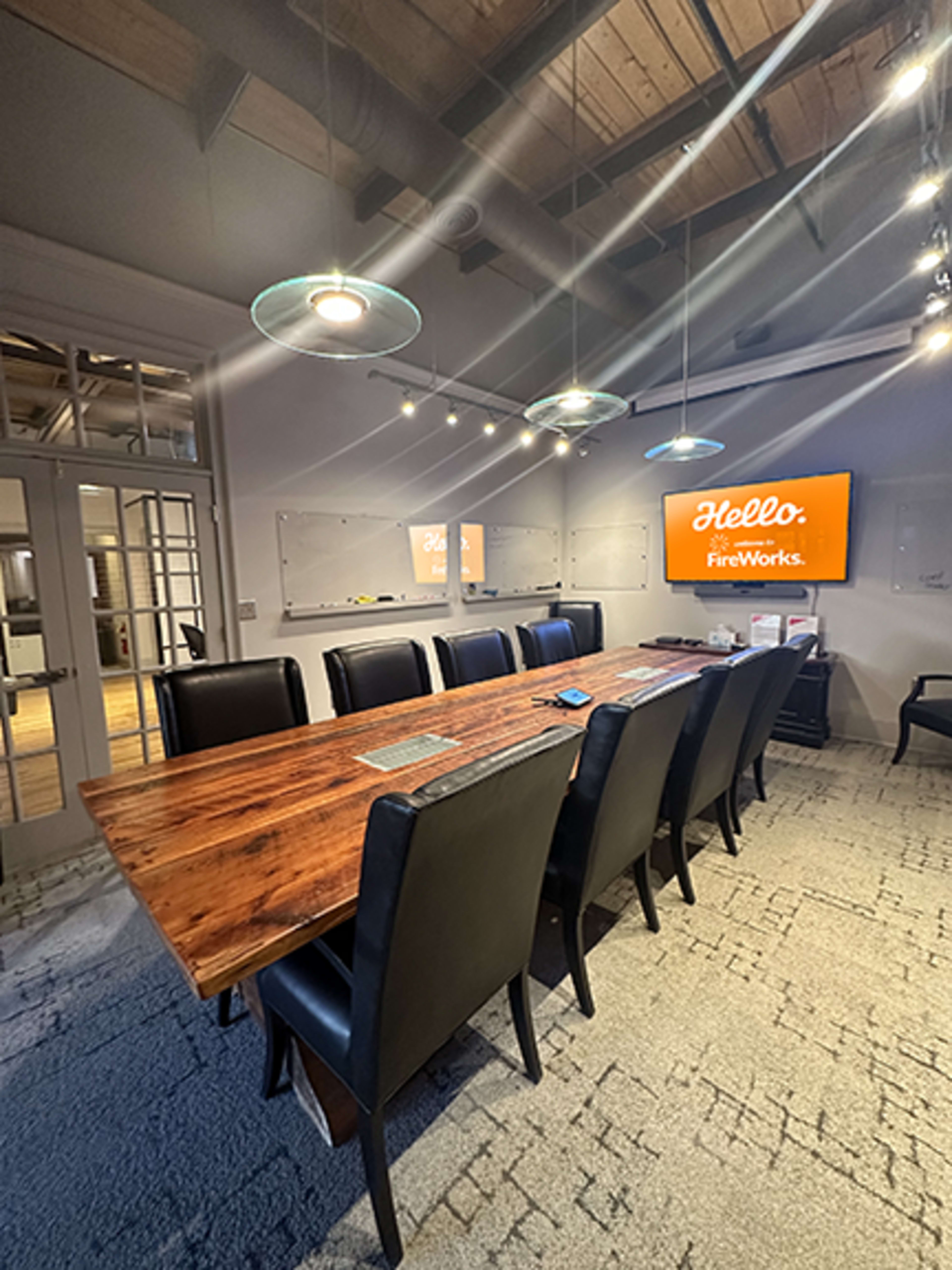 A modern conference room features a large wooden table surrounded by black chairs, with a screen displaying "Hello, FireWorks" on the wall.