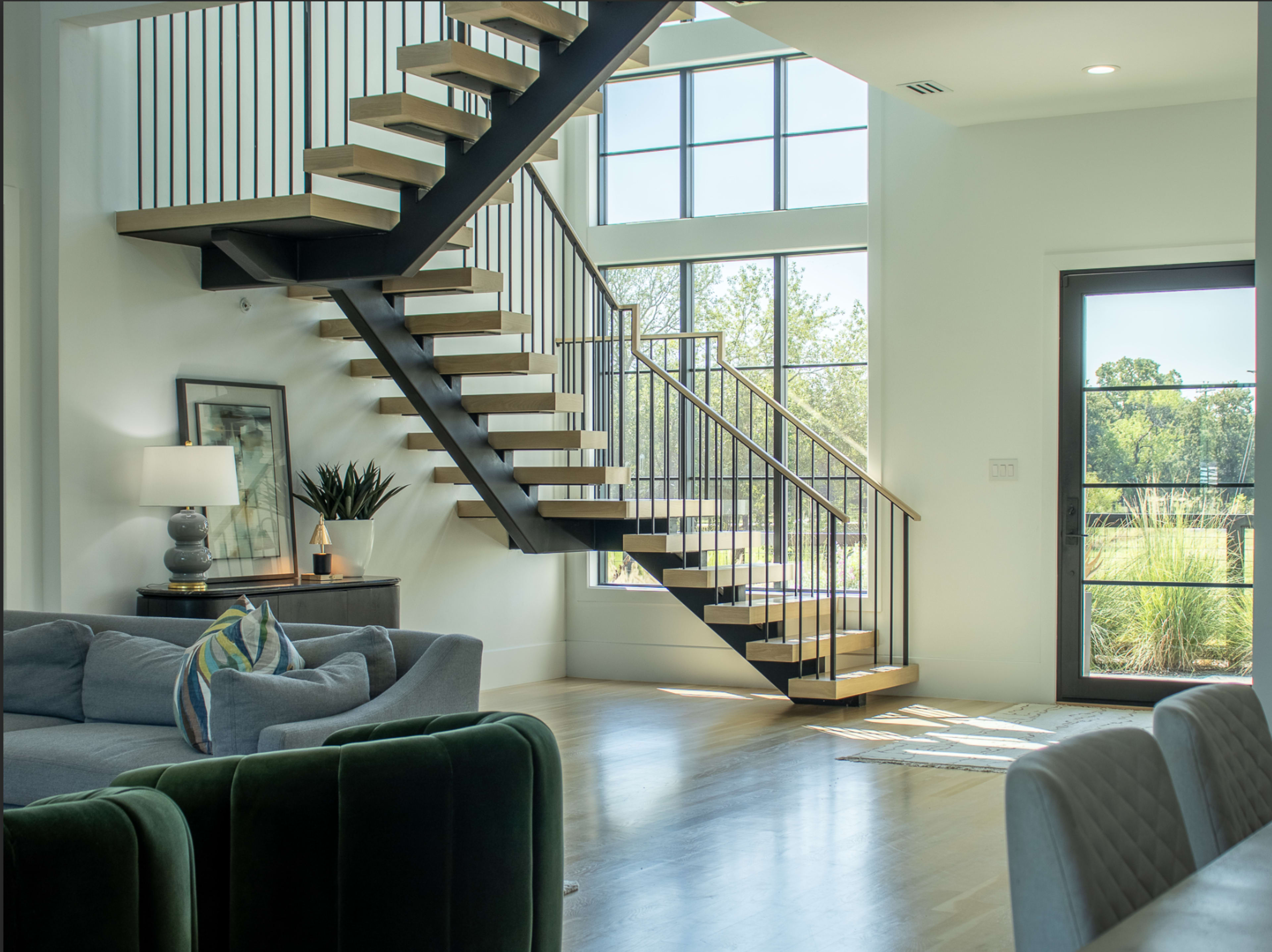 The image shows a modern interior with a staircase, large windows, and a living area featuring a gray sofa and a green velvet chair.