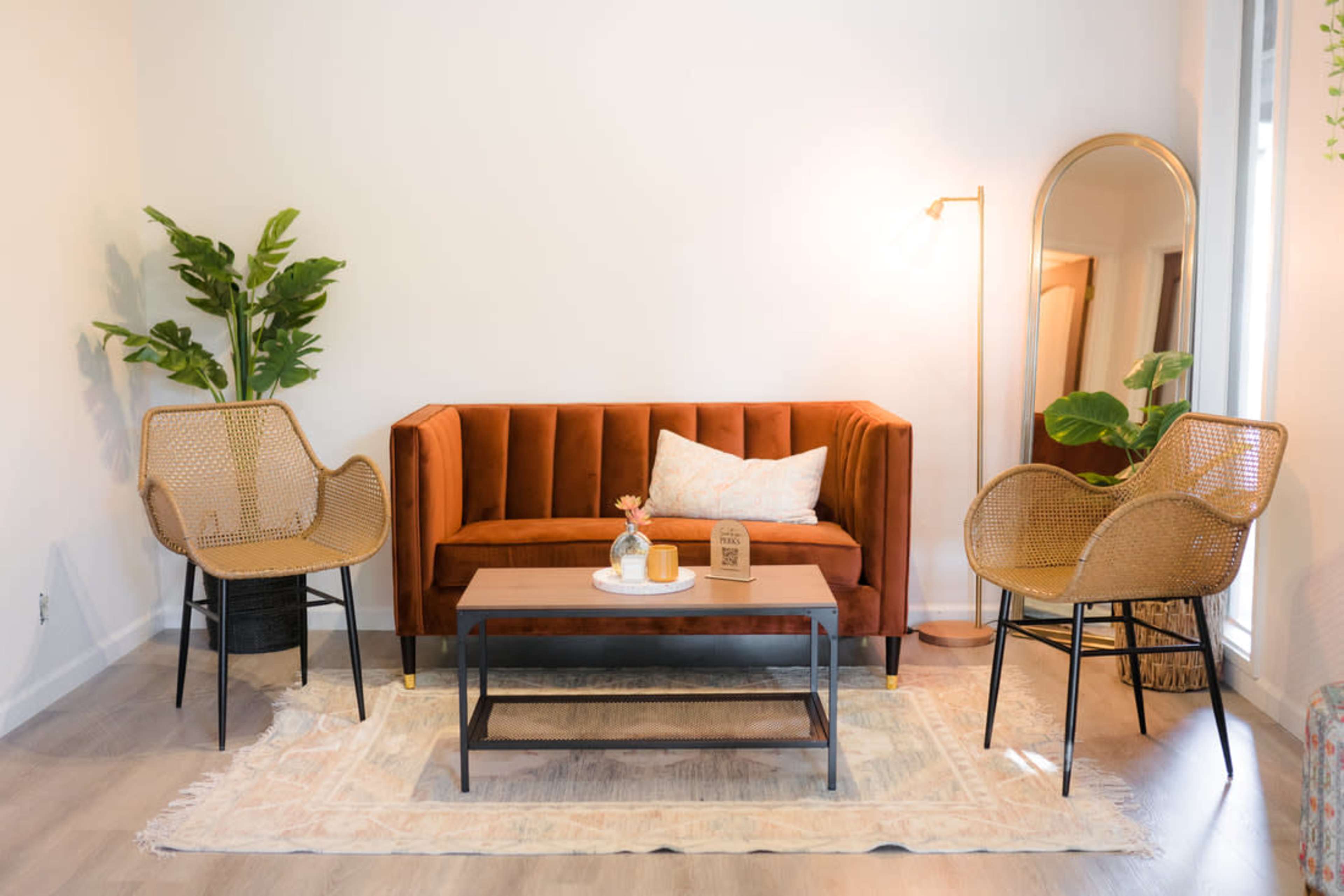 A modern living room with an orange velvet sofa, two woven chairs, a wooden coffee table, a decorative vase, and a floor lamp, set against a light-colored wall.