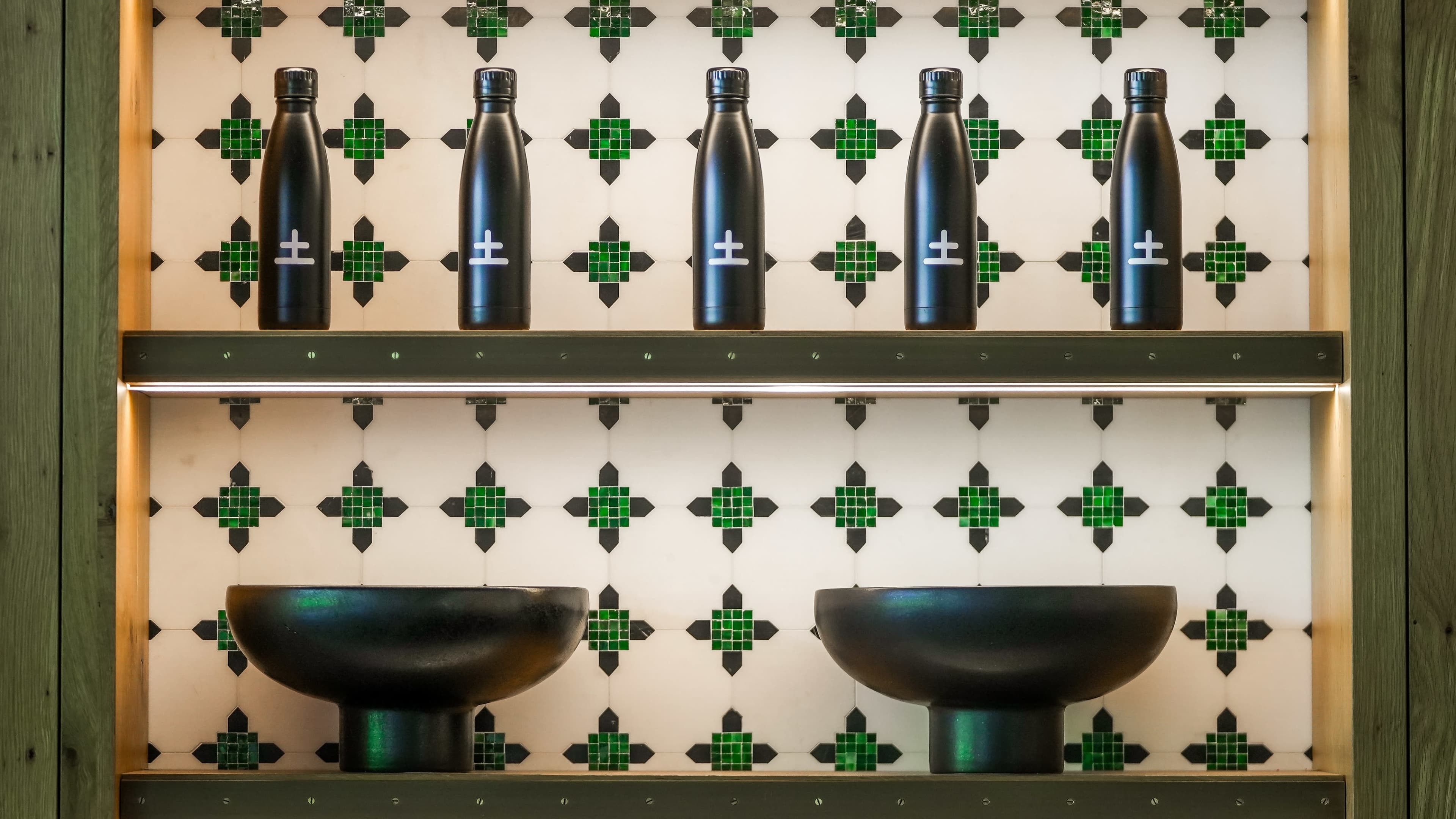 The image features a wall display with six black bottles arranged on a shelf above two dark bowls, set against a tiled background with a green and white geometric pattern.