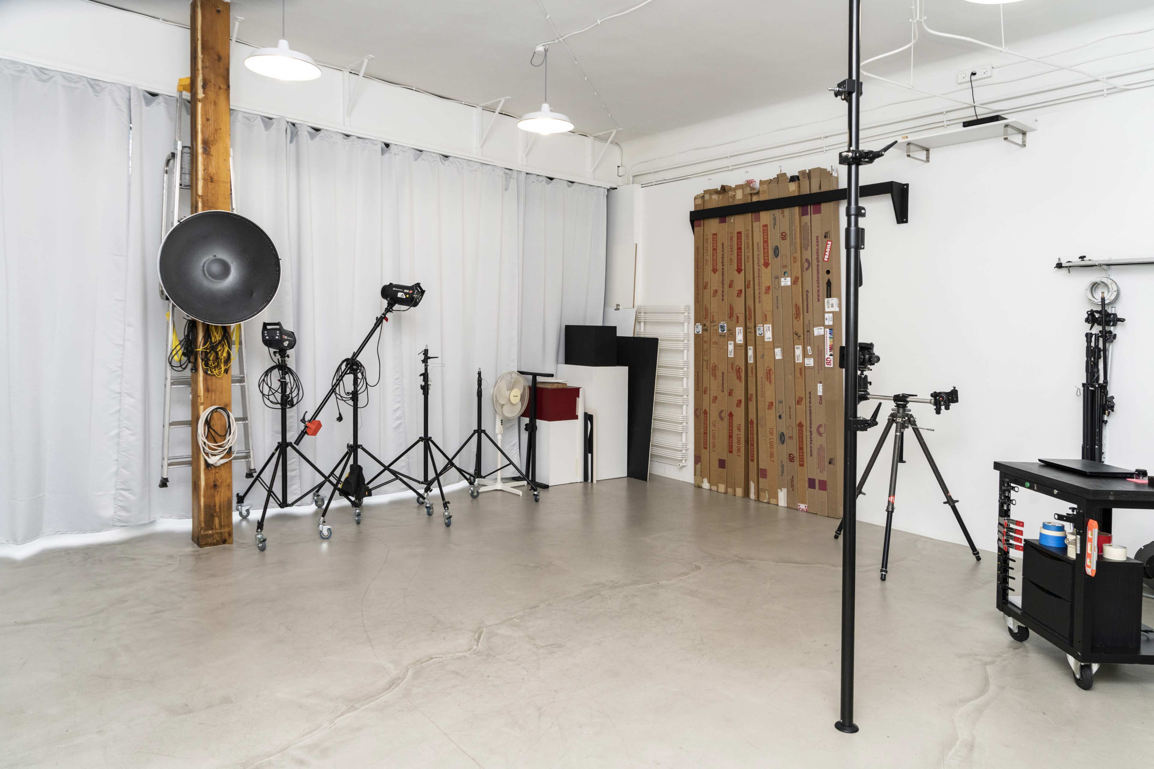 The image shows a photography studio with various lighting equipment, tripods, and a backdrop against a white wall.