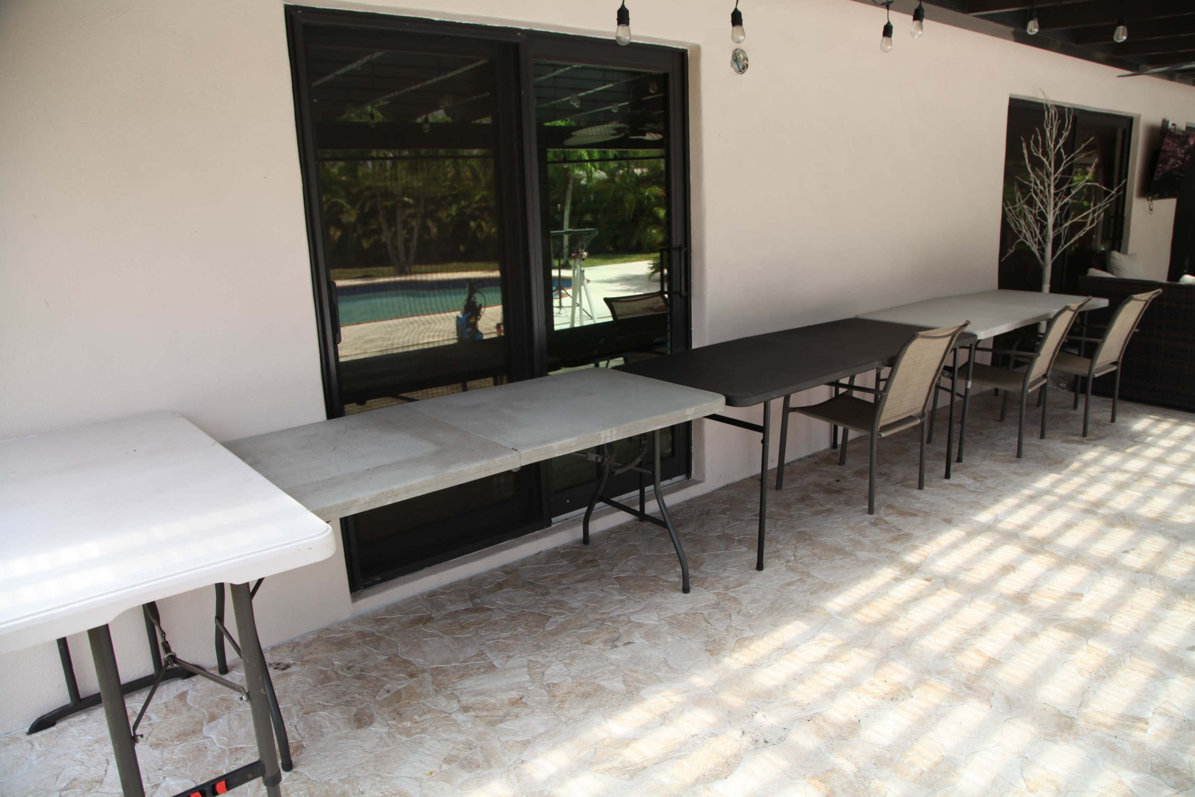 A row of folding tables is lined up on a tiled patio adjacent to large windows overlooking a swimming pool.