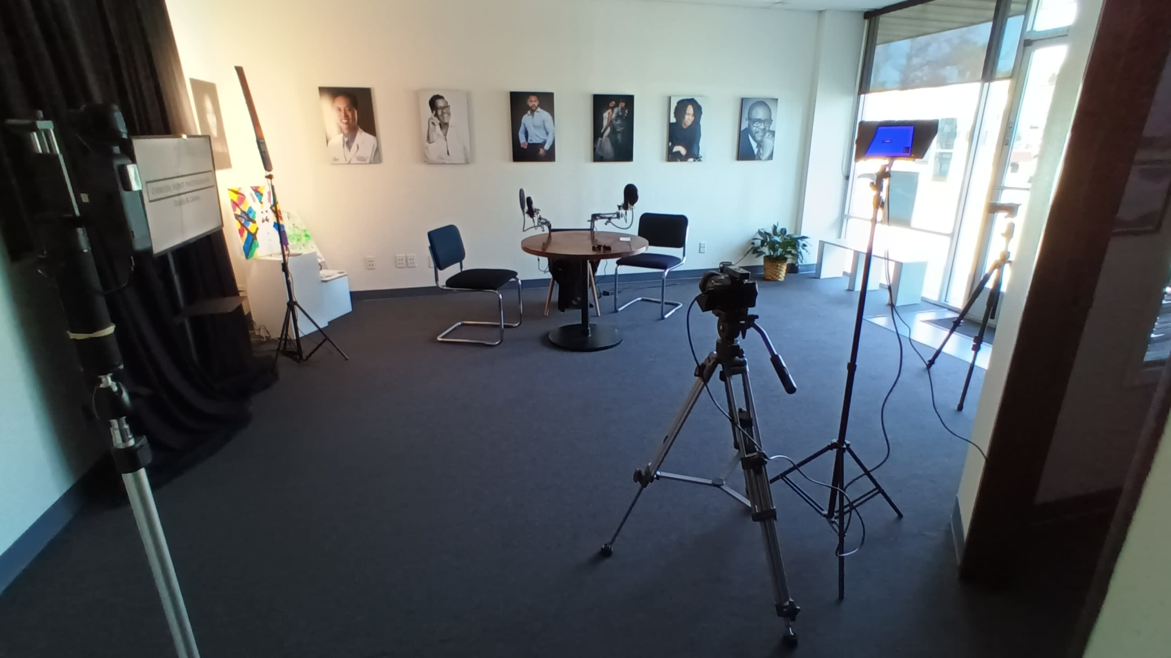 The image shows a recording studio featuring a circular table and chairs, with microphones set up, a camera on a tripod, and several framed portraits on the wall.