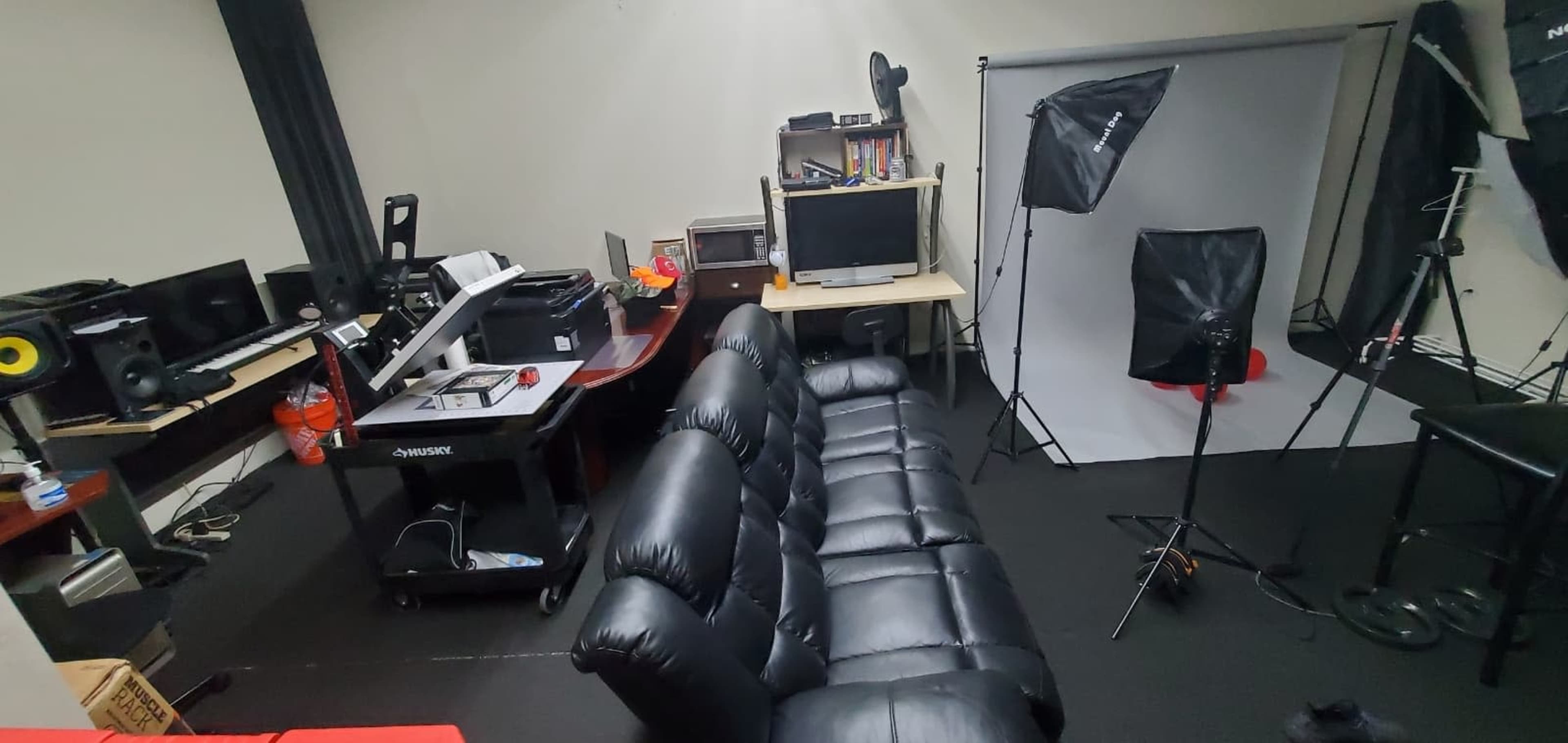 A recording studio setup with a black leather couch, various audio equipment, a desk with a computer, and lighting equipment positioned in the corner.