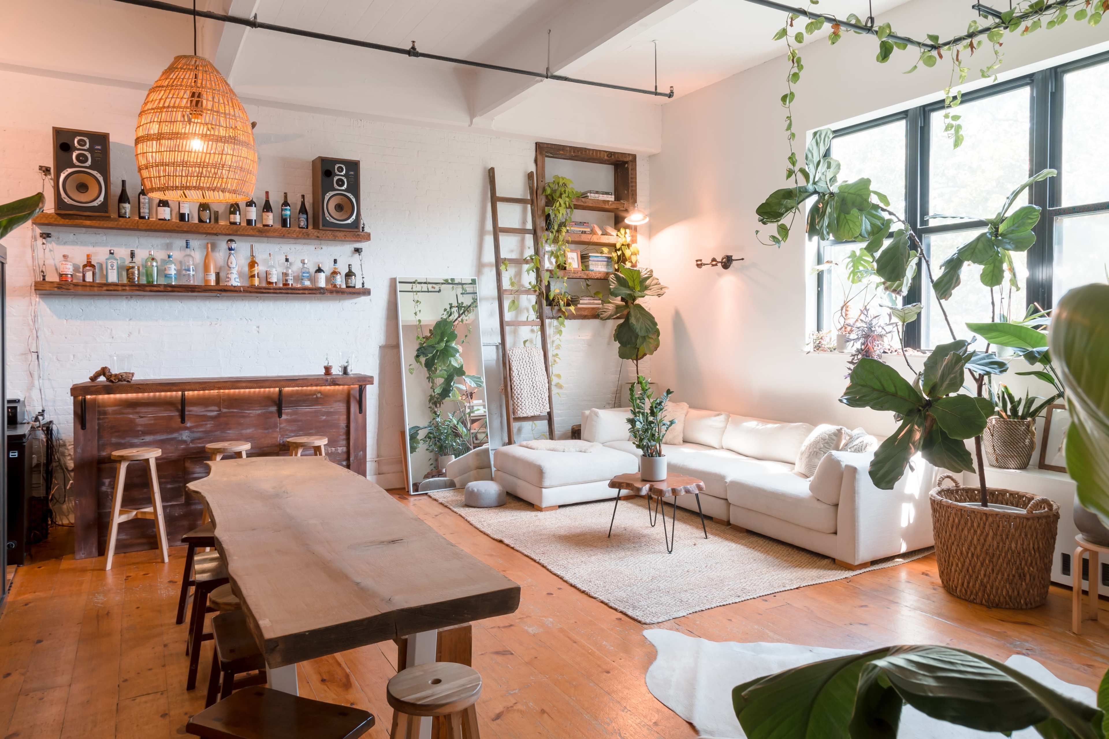 A spacious, well-lit room featuring a bar area, a large dining table, a white sofa, numerous houseplants, and wooden accents.
