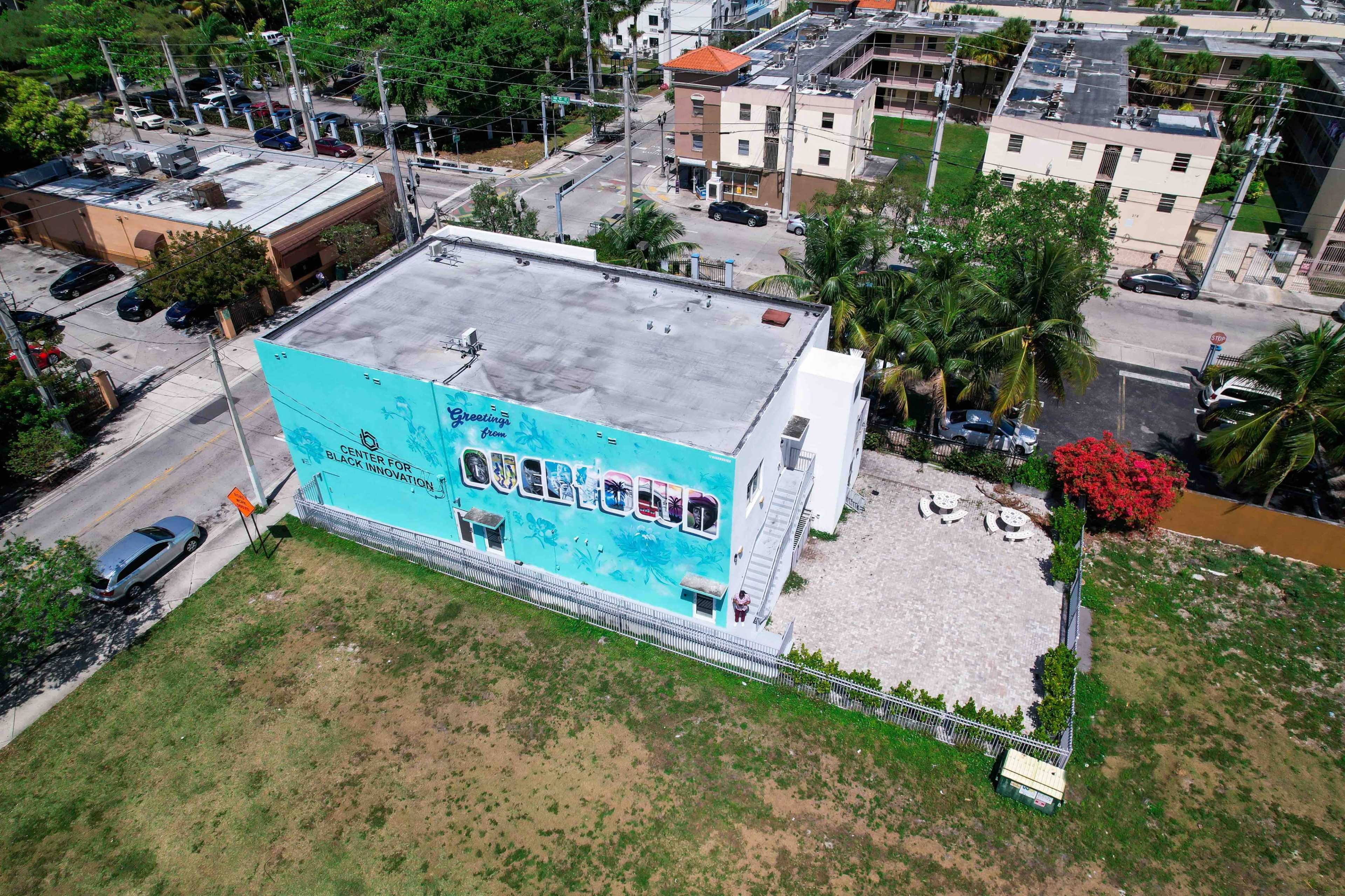 An aerial view shows a building with a colorful mural and a landscaped outdoor area surrounded by residential structures and streets.