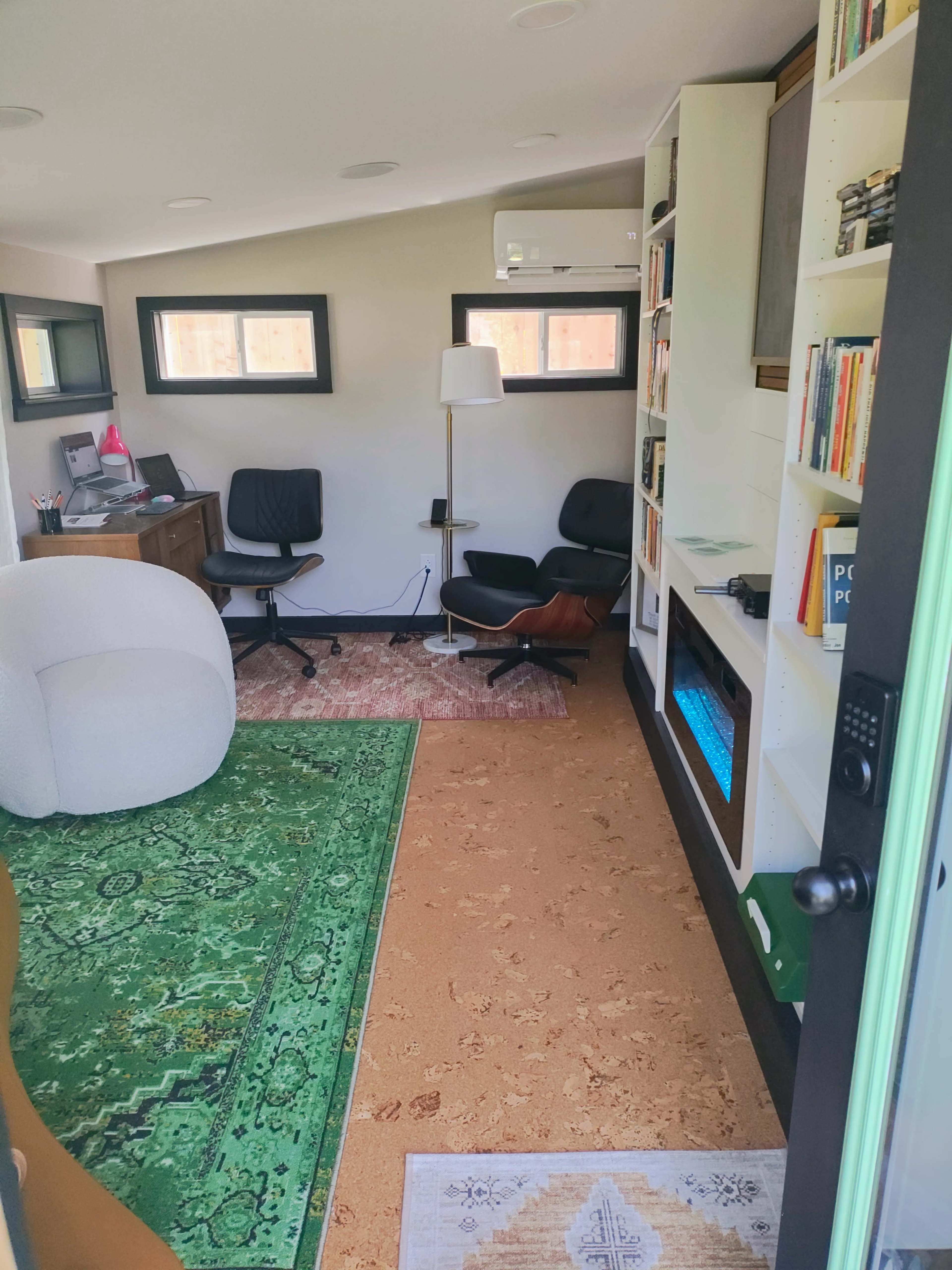 The image shows a small office with a green rug, a bookshelf filled with books, a desk with chairs, and a window allowing natural light.