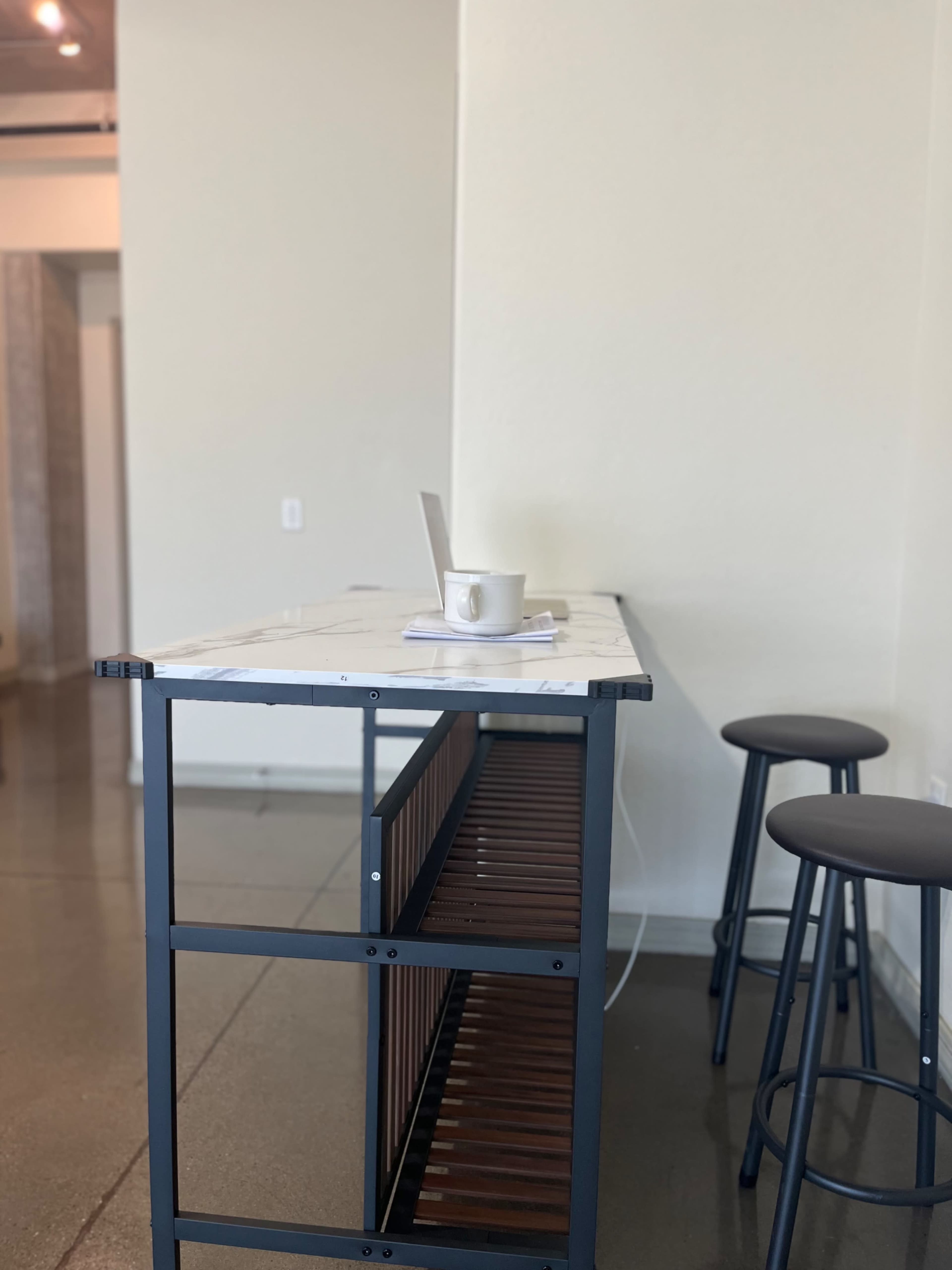 A minimalist workspace features a marble-topped bar with a laptop and a cup on it, accompanied by two metal stools.
