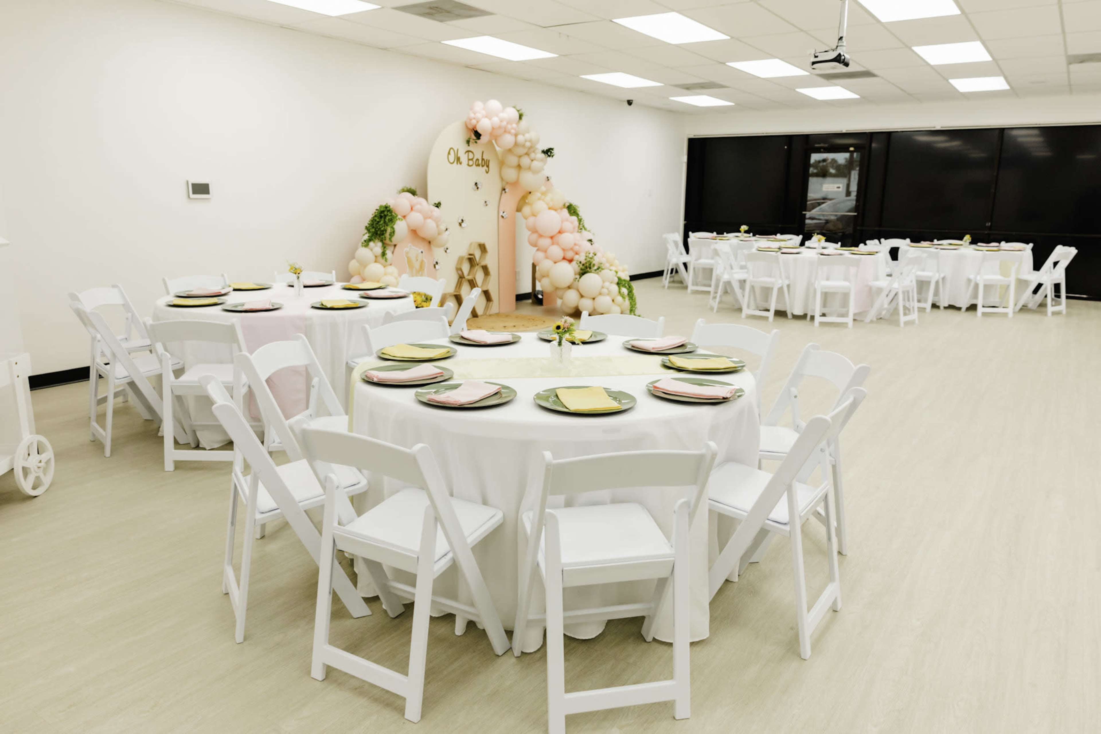 The image shows a decorated event space with round tables covered in white tablecloths and set with plates, surrounded by a backdrop of balloons and greenery.