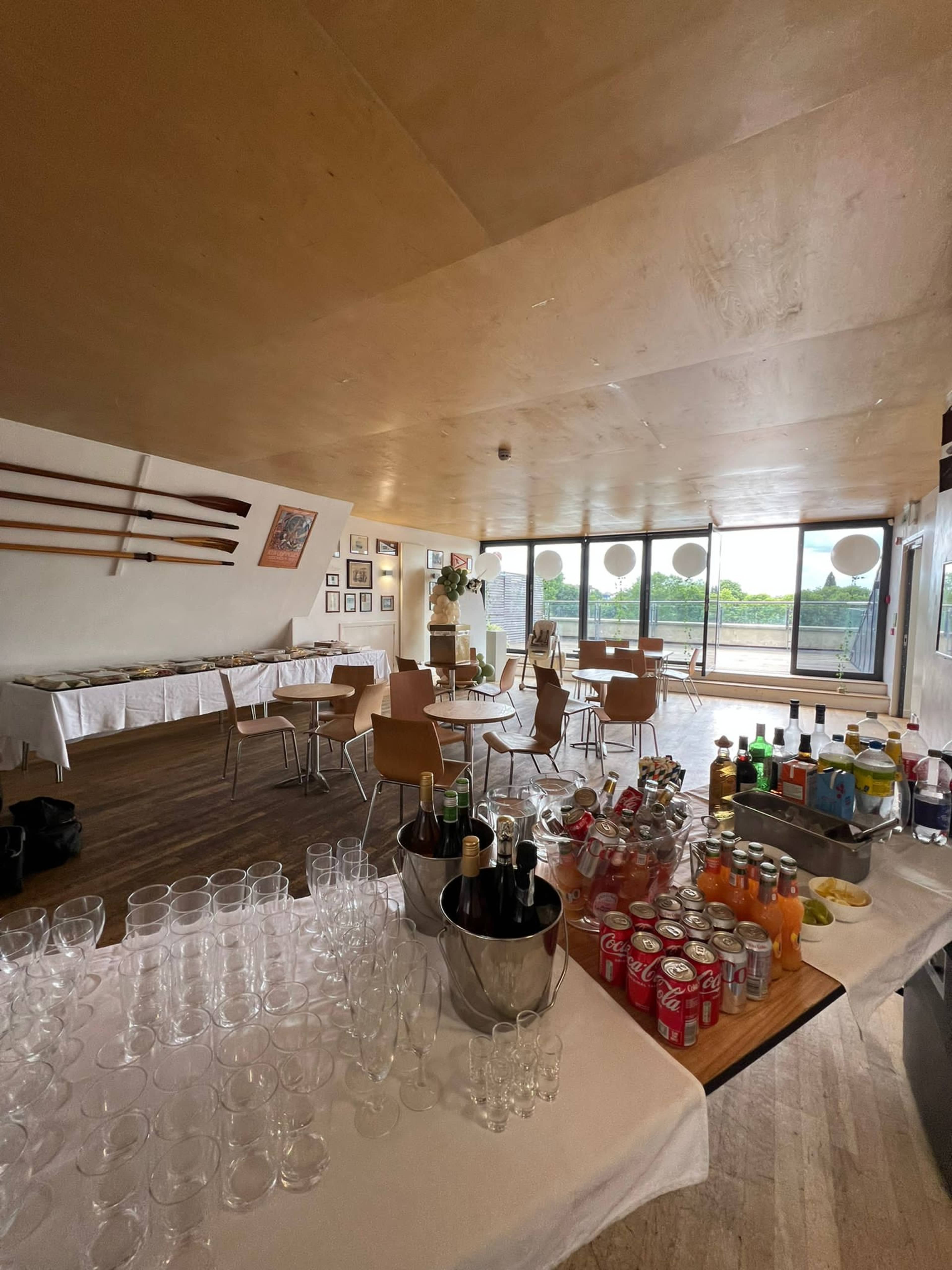 The image shows an indoor event space with tables set for refreshments, a variety of drinks displayed, and large windows overlooking a landscape.