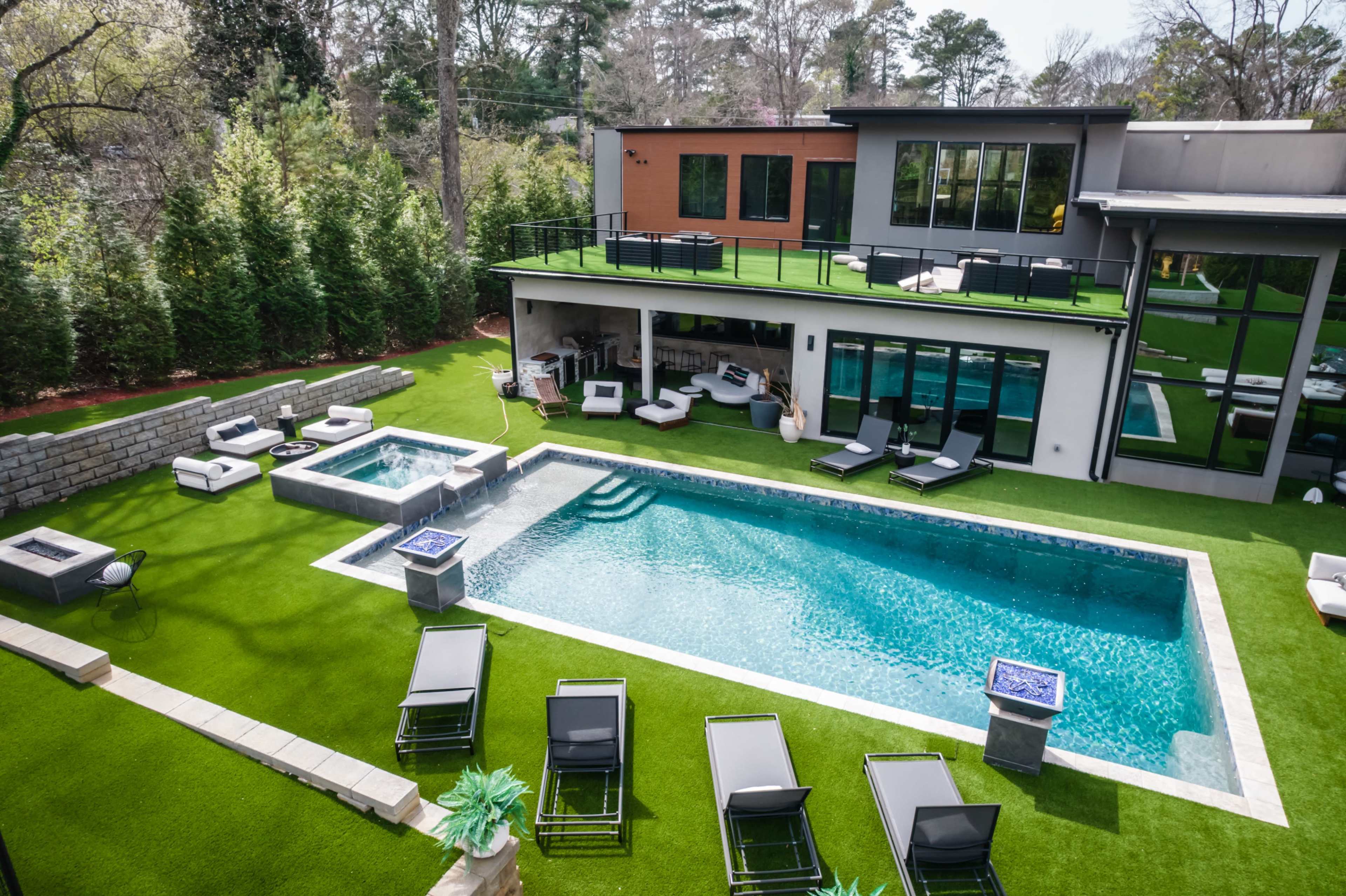 The image shows a modern backyard featuring a swimming pool, hot tub, and multiple seating areas surrounded by greenery.