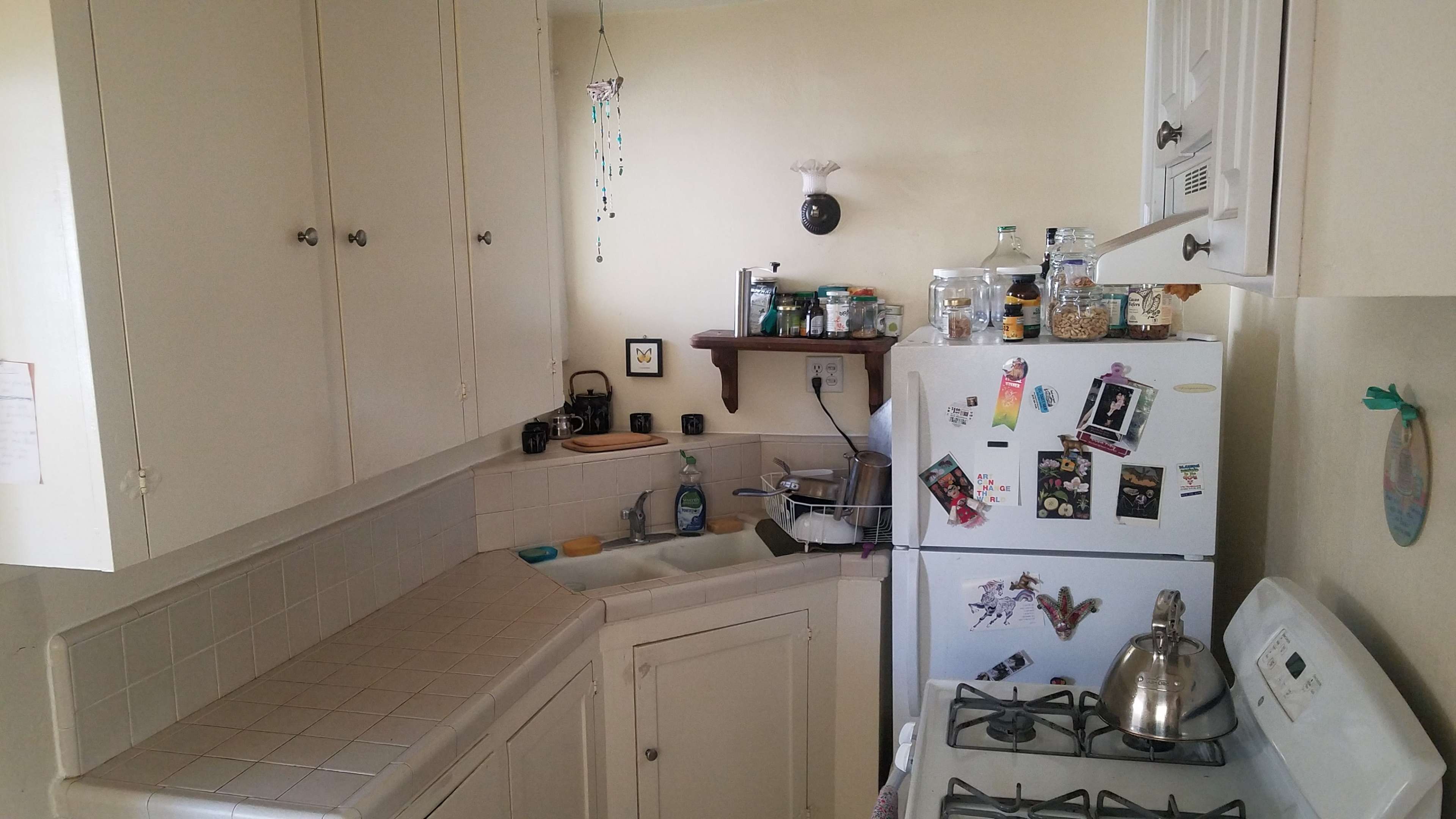 The image shows a small kitchen with white cabinets, a sink, a stove, and a refrigerator adorned with various stickers.
