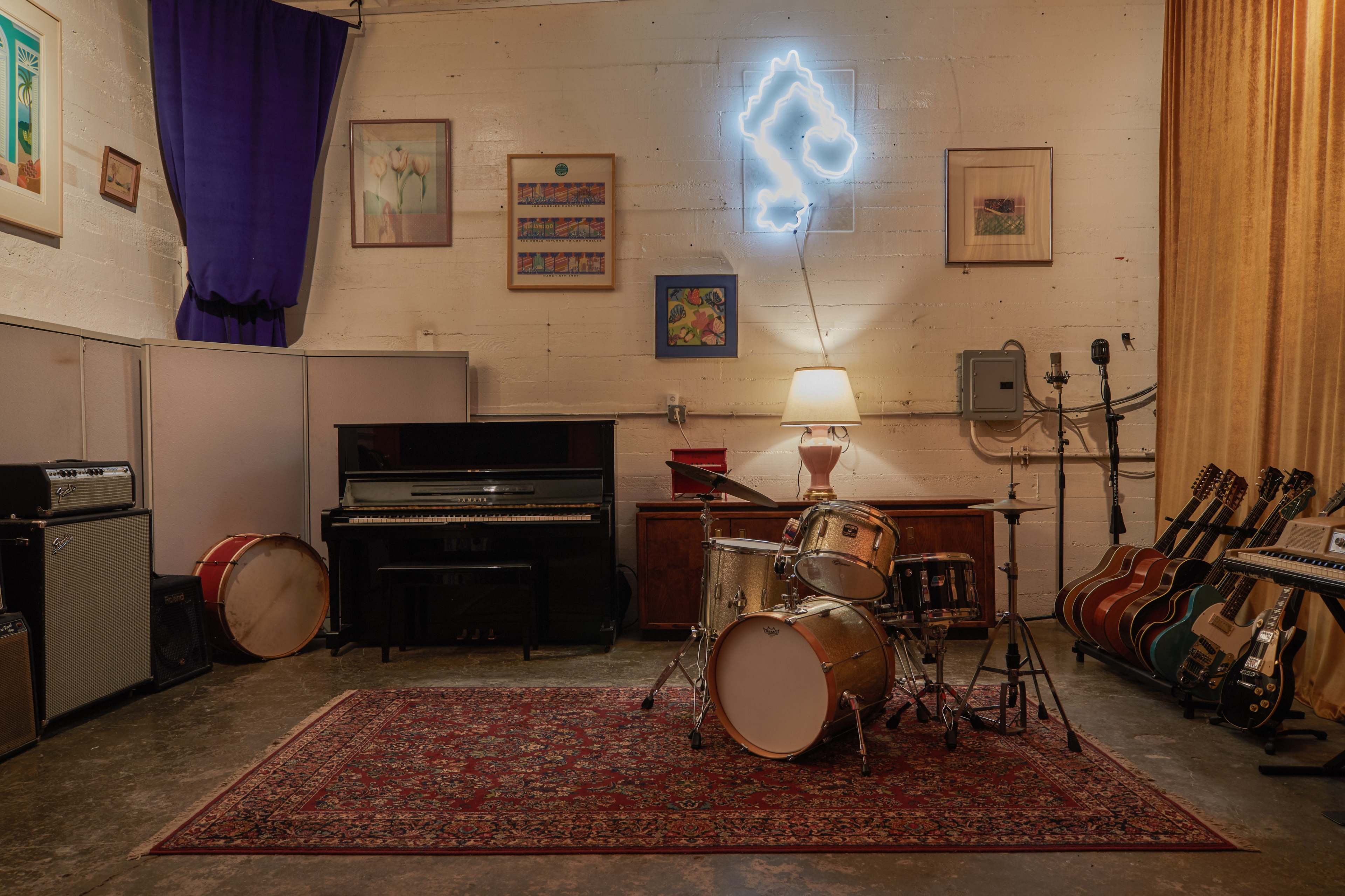 A music rehearsal space features a drum set, a piano, amplifiers, and various guitars, all arranged around a colorful rug under soft lighting.