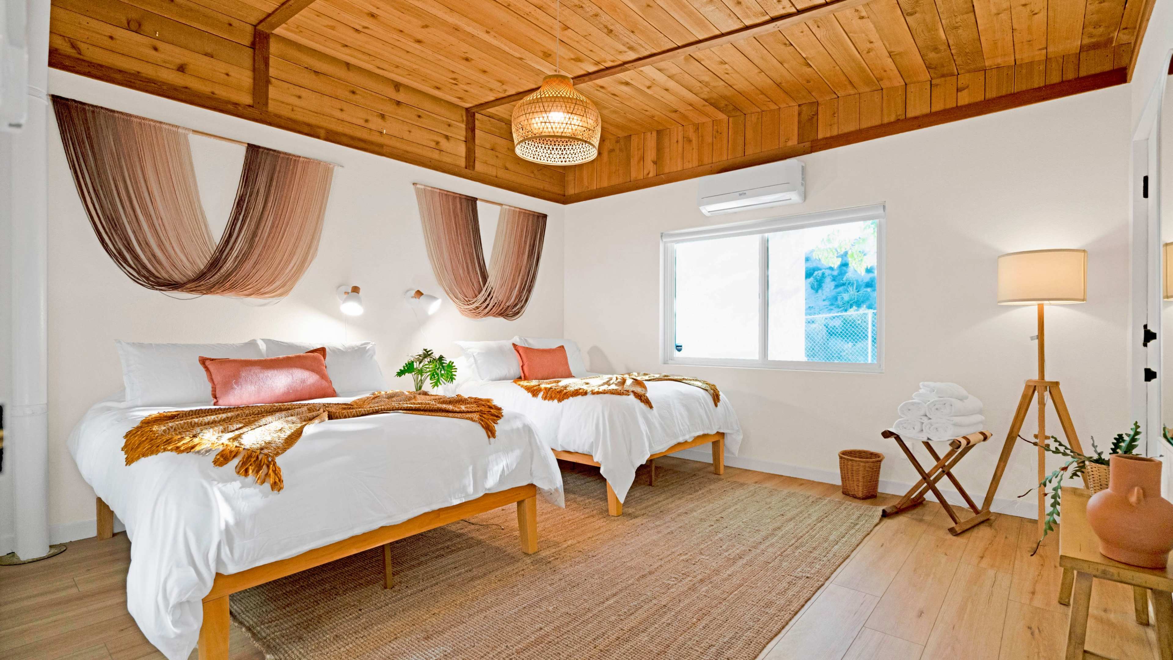The image shows a bright bedroom with two beds, wooden ceilings, and decorative wall hangings, featuring a window that lets in natural light.
