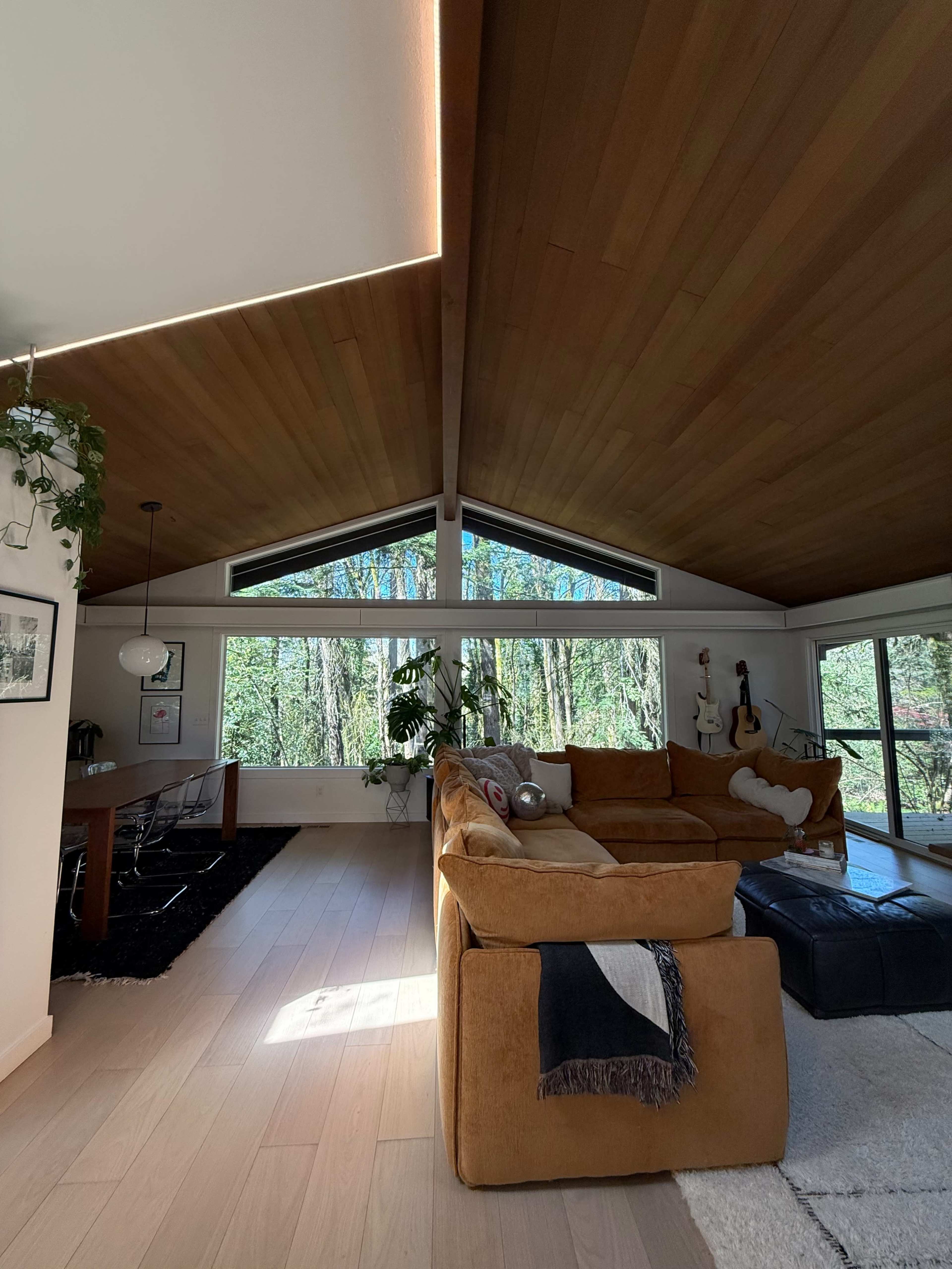 The image shows a spacious living room with a wooden ceiling, large windows overlooking a wooded area, and a cozy sectional sofa.