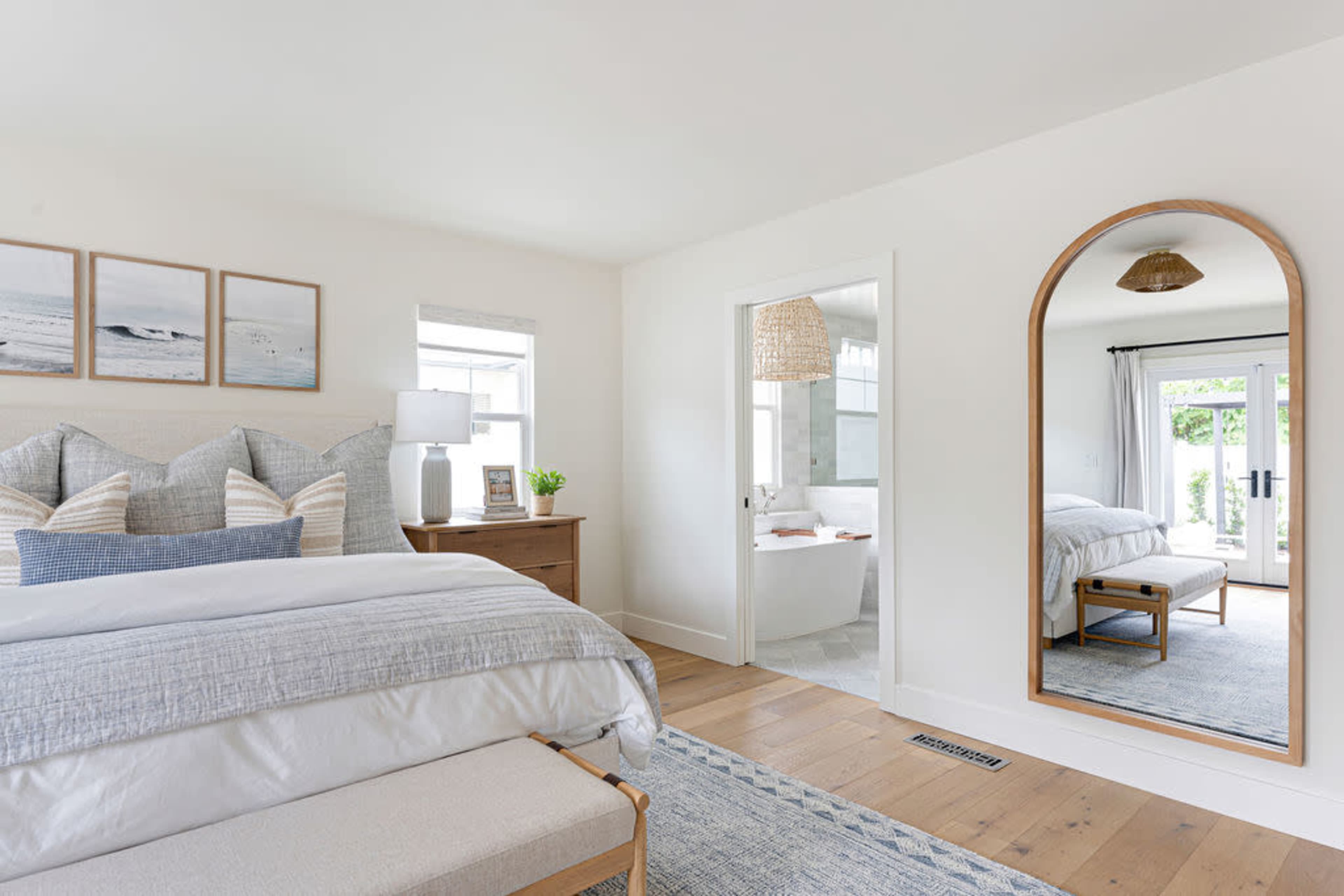 A modern bedroom features a large bed with decorative pillows, a wooden dresser, and a round mirror, with a view of an adjacent bathroom through an open door.