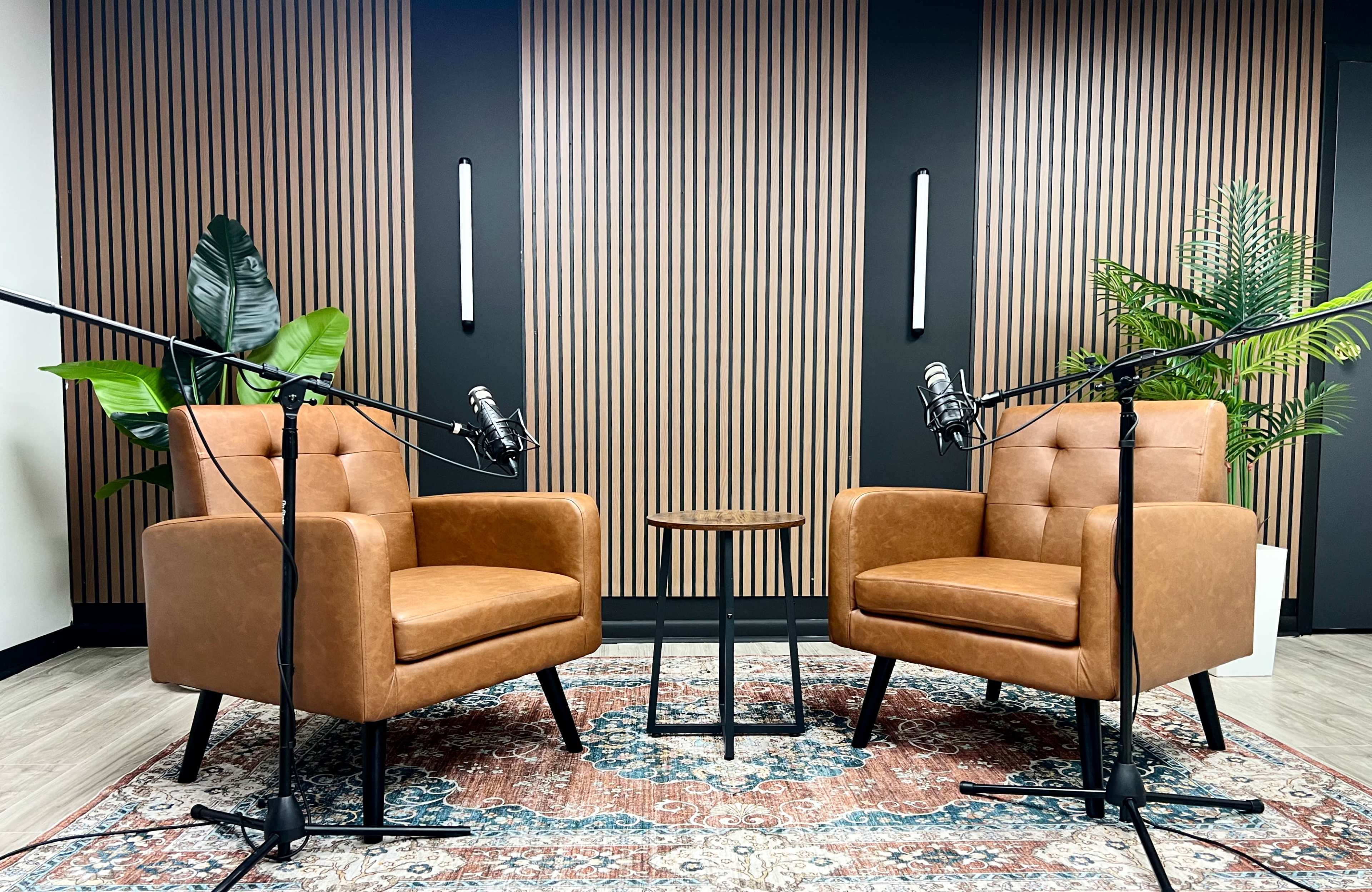 The image shows a podcasting setup with two brown armchairs facing each other, a small table in between, and microphones mounted on stands.