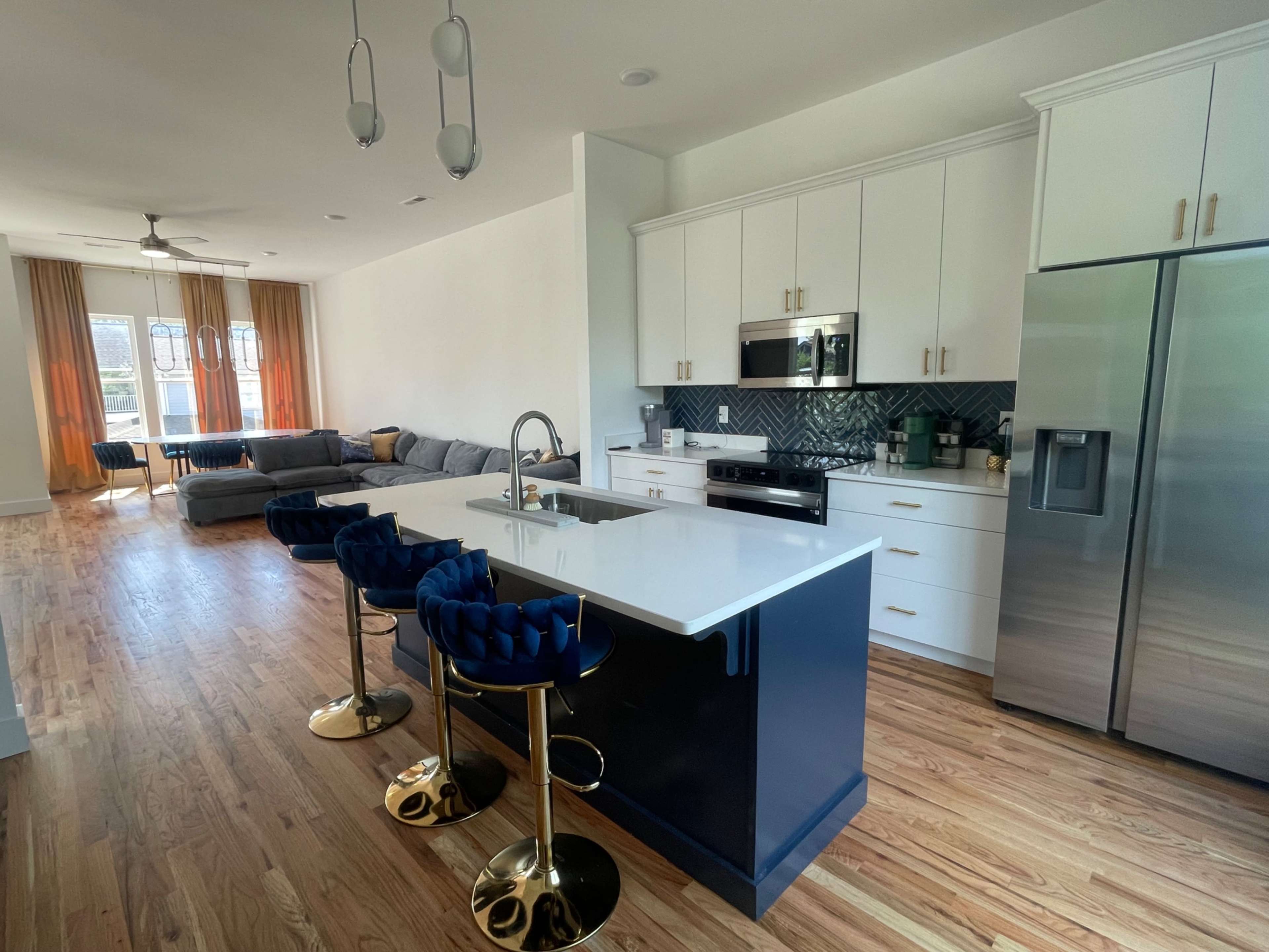 The image shows a modern kitchen with a large island featuring blue and white cabinetry, three gold bar stools, and an open living area with a gray couch and large windows.