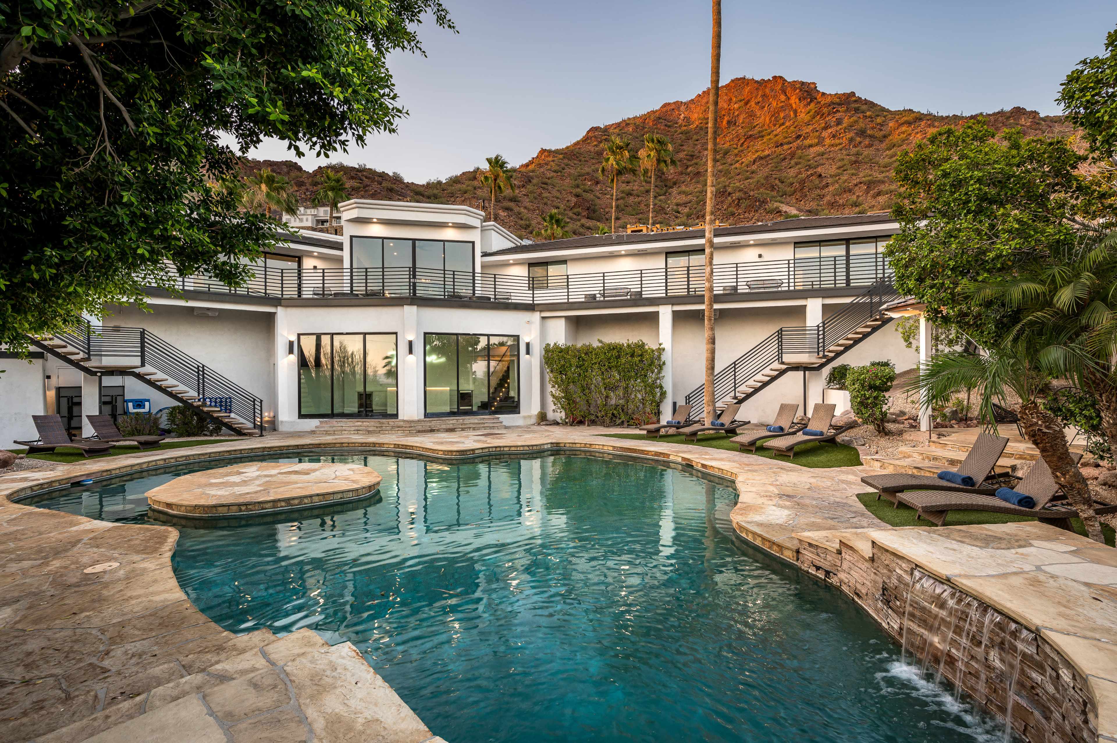 A modern home with a swimming pool surrounded by stone decking and palm trees, set against a mountain backdrop.