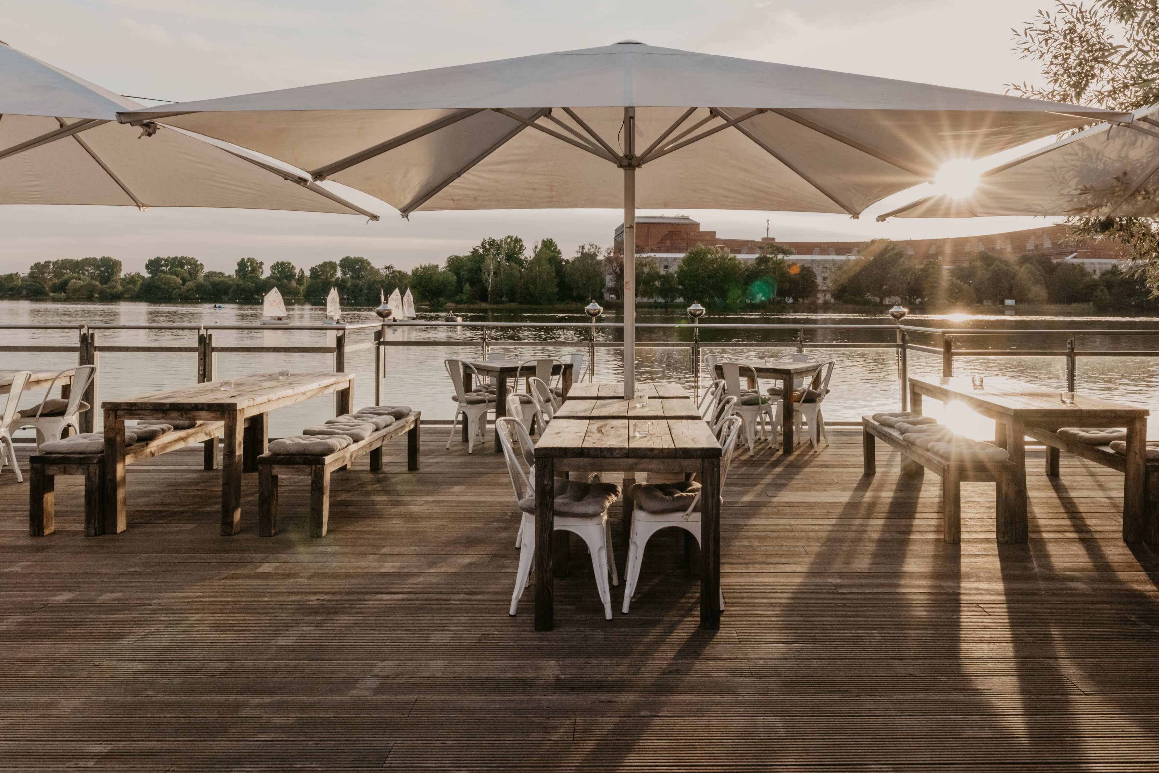 The image shows an outdoor dining area with wooden tables and white chairs, set by a river under large umbrellas, with a sunset casting long shadows.