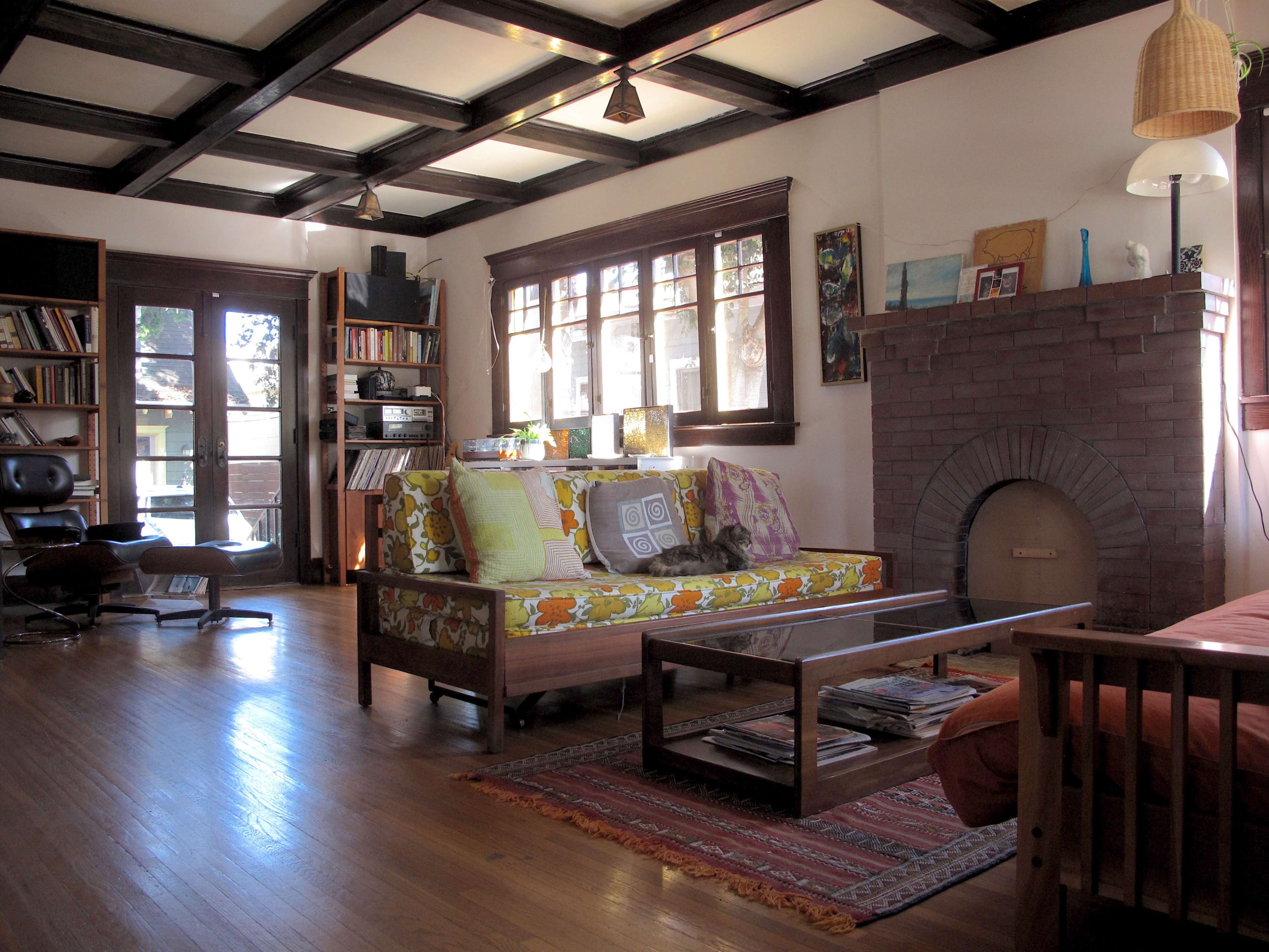 A cozy living room features a patterned sofa, a wooden coffee table with magazines, shelves filled with books, and a brick fireplace under a coffered ceiling.
