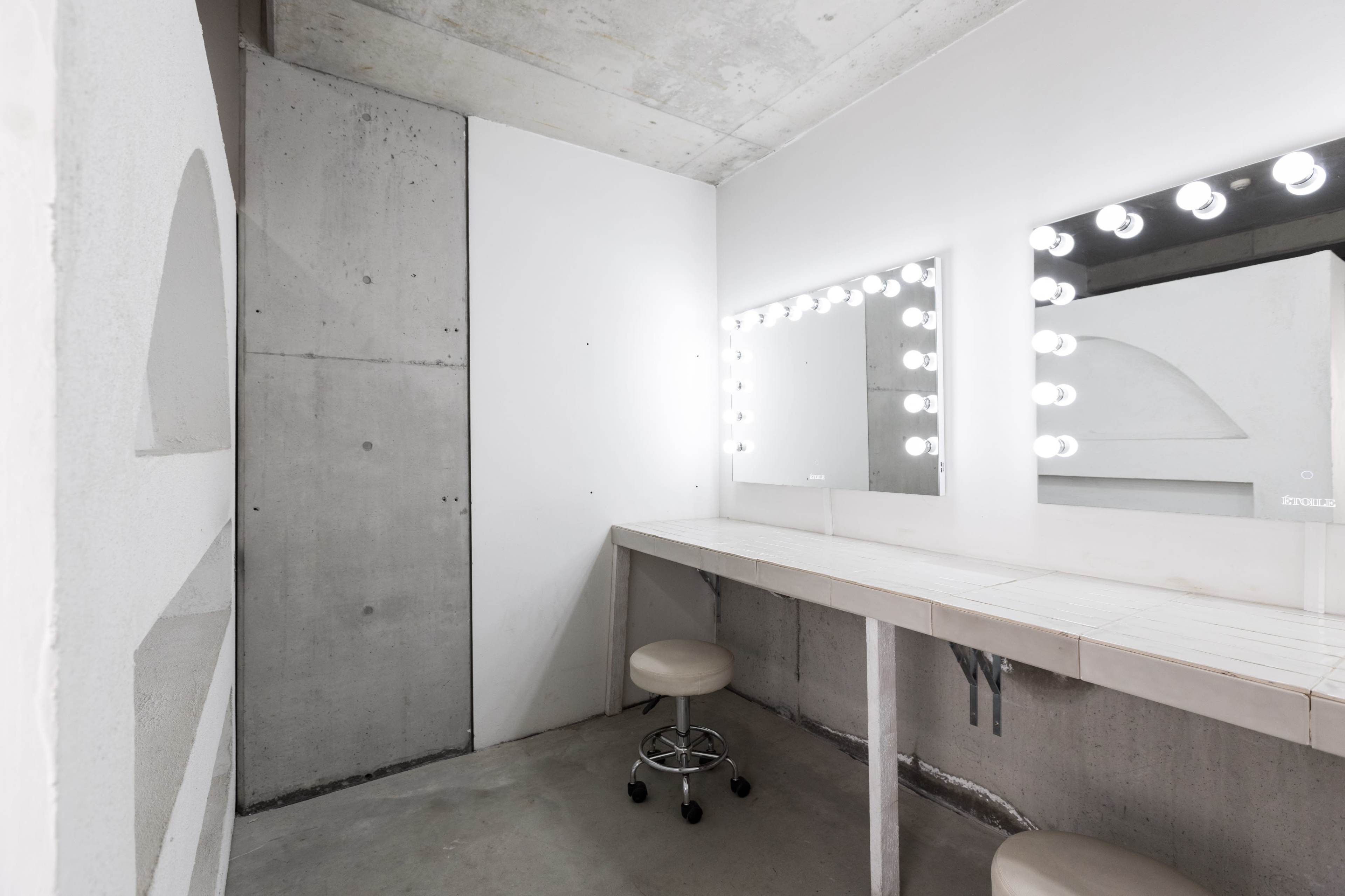A minimalist dressing room with a white vanity table, two illuminated mirrors, and a swivel stool against a concrete wall.