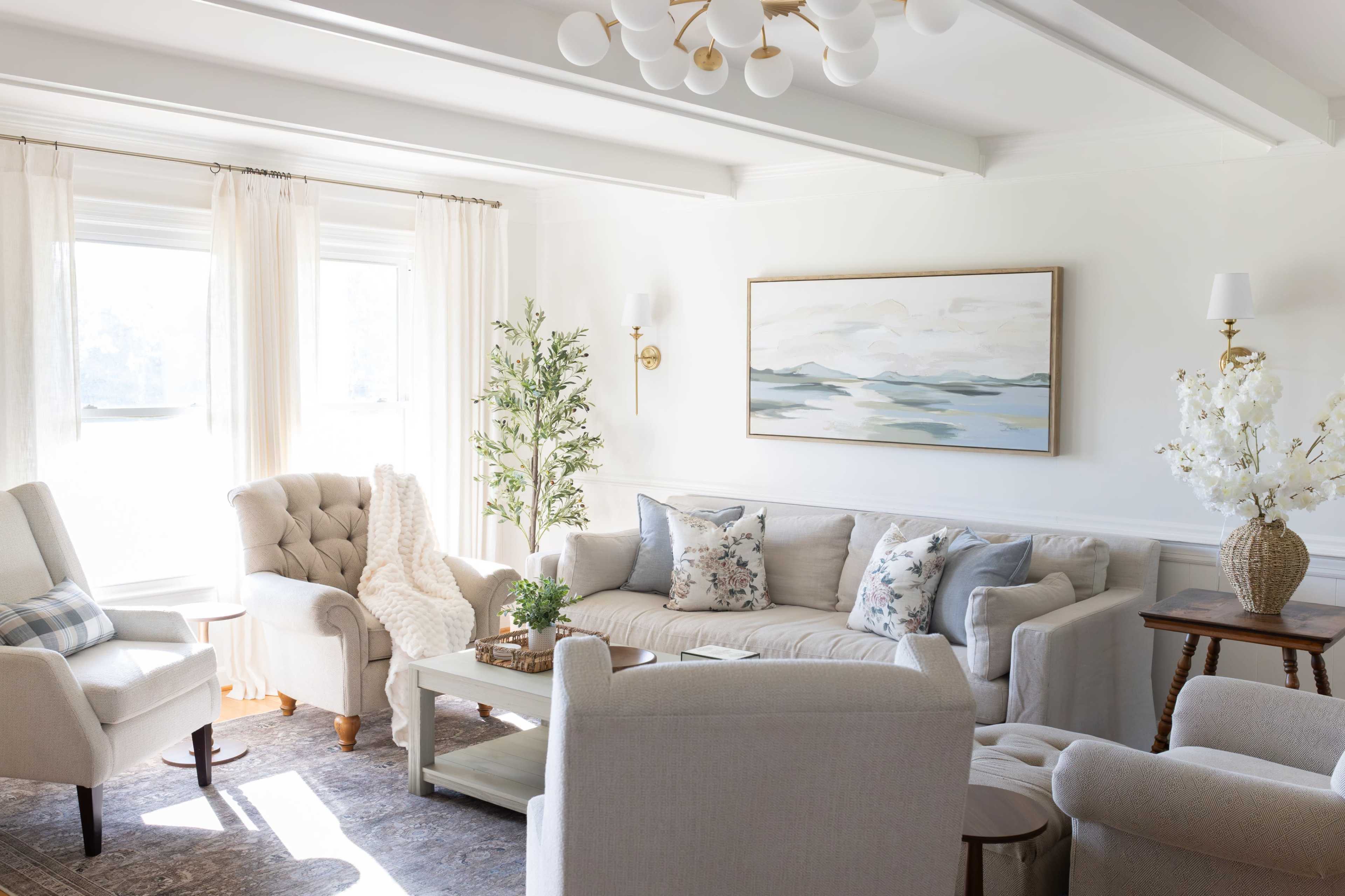 The image shows a bright, cozy living room featuring a sofa with decorative pillows, two armchairs, a coffee table, and a large painting on the wall.