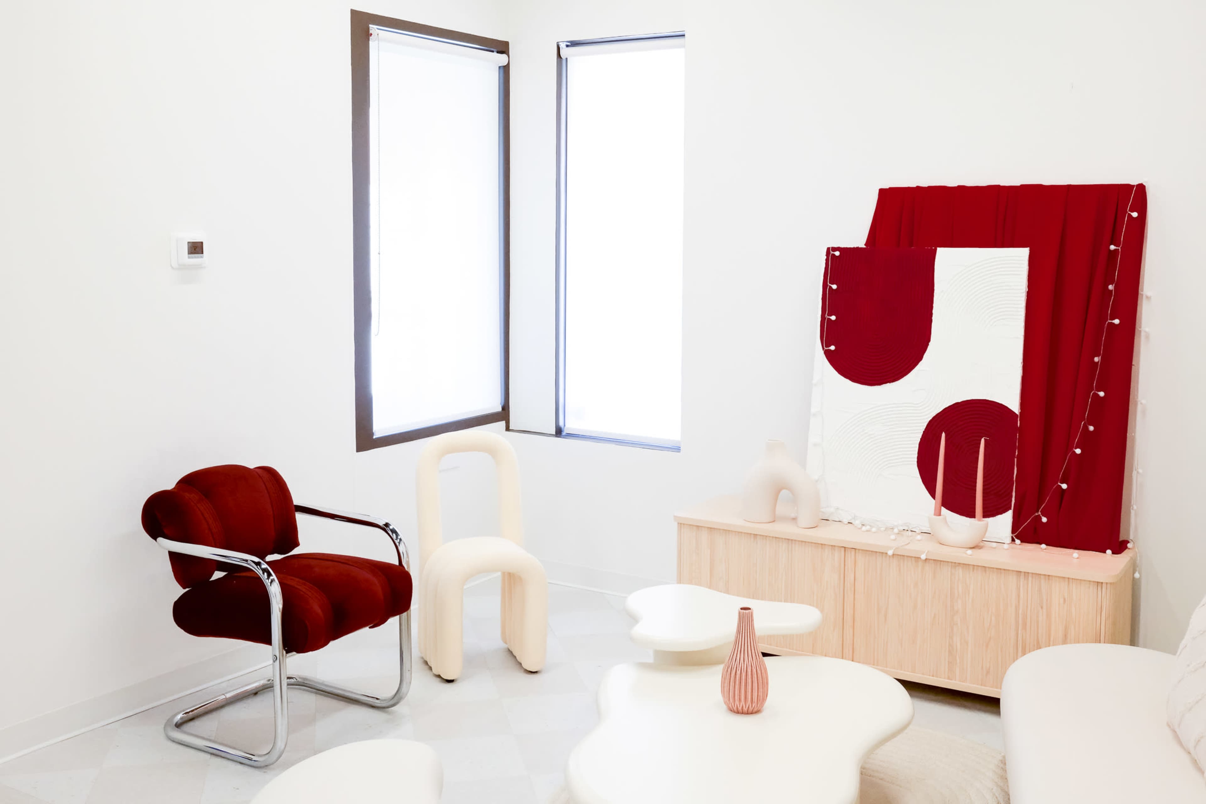 A modern interior with a red chair, a light wooden cabinet, abstract art on the wall, and large windows letting in natural light.